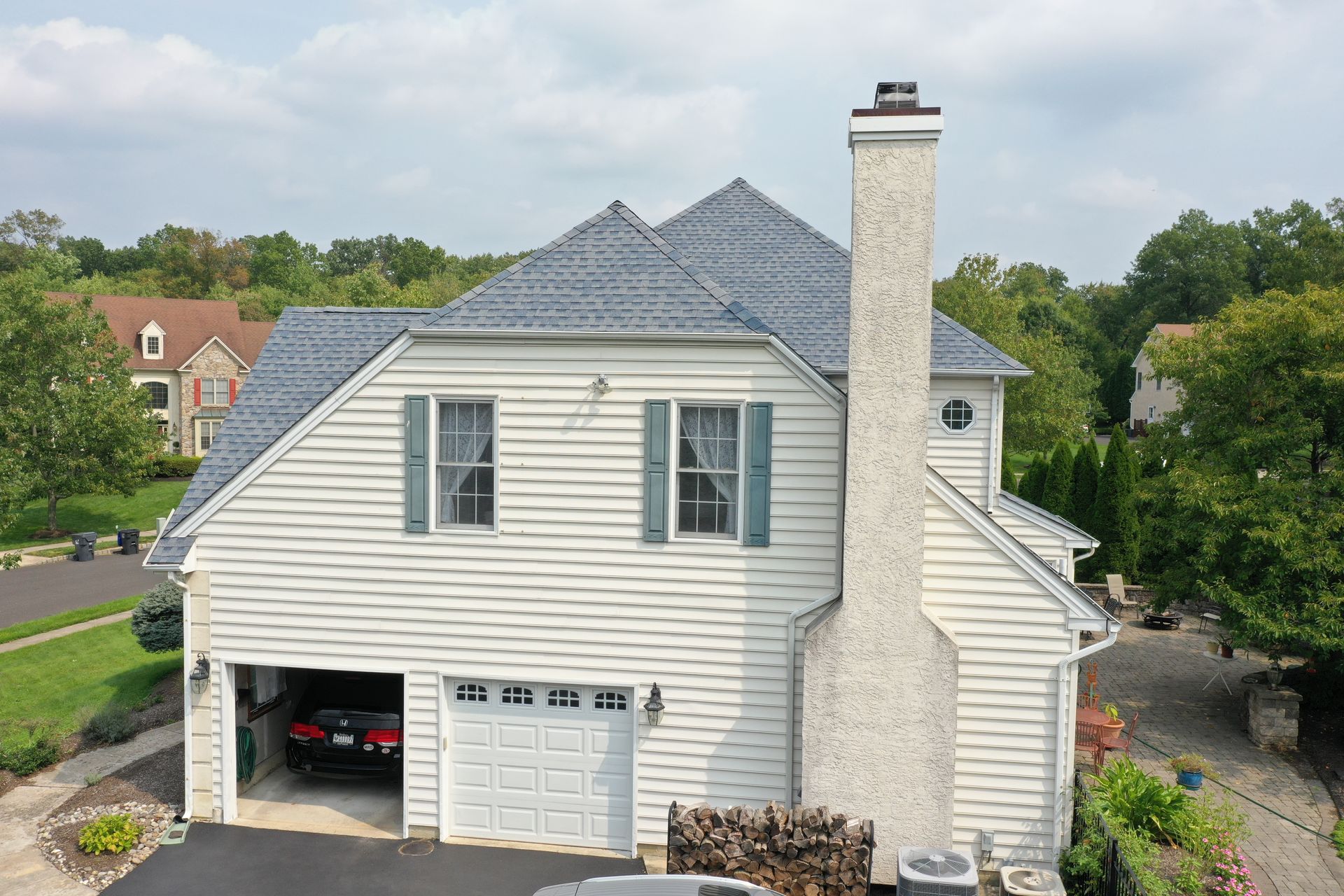 White house with a gray roof and a tall chimney. Car parked in the garage, trees in the background.