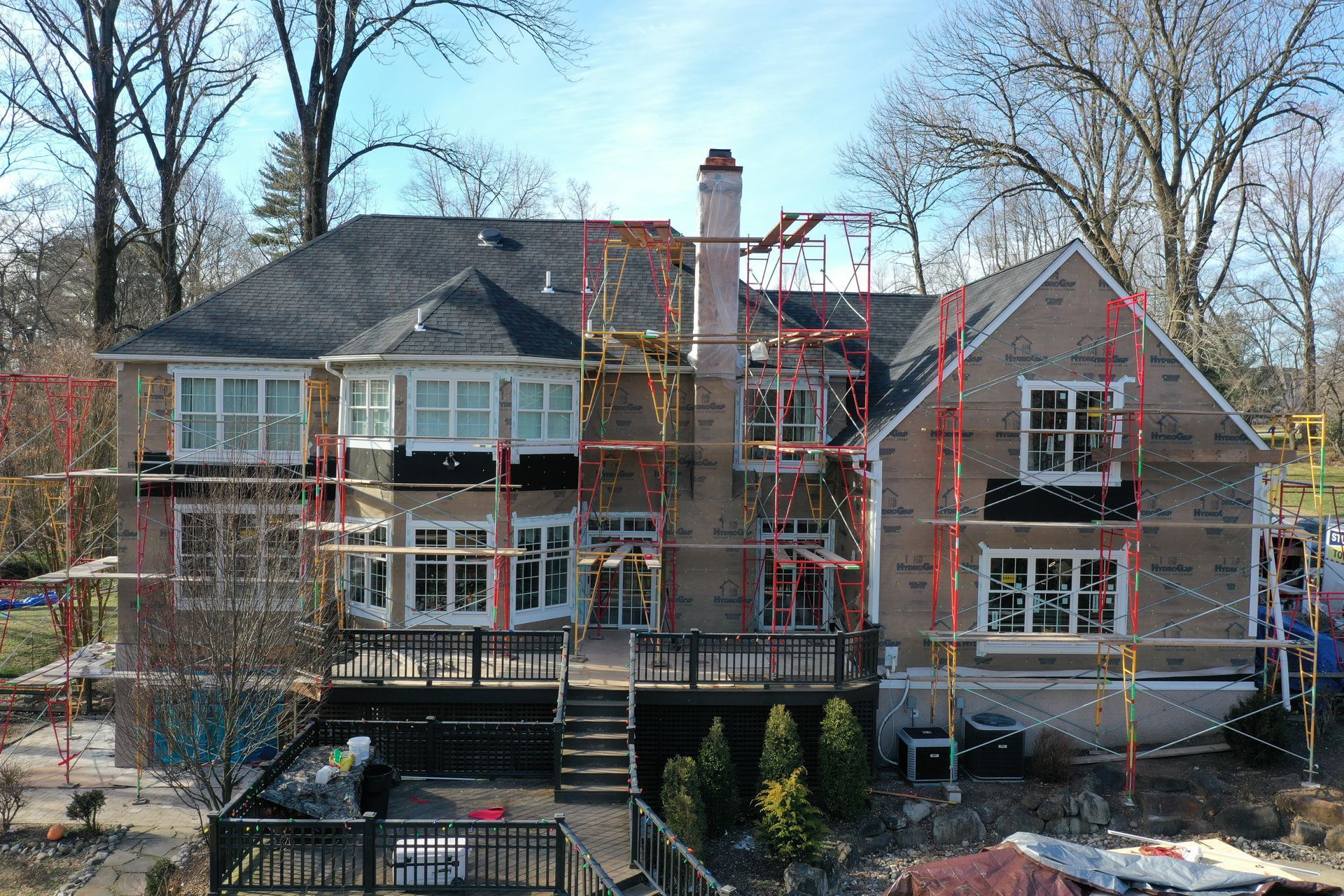 Back of a two-story brick house with scaffolding during renovation.  Deck, trees, and sky are visible.