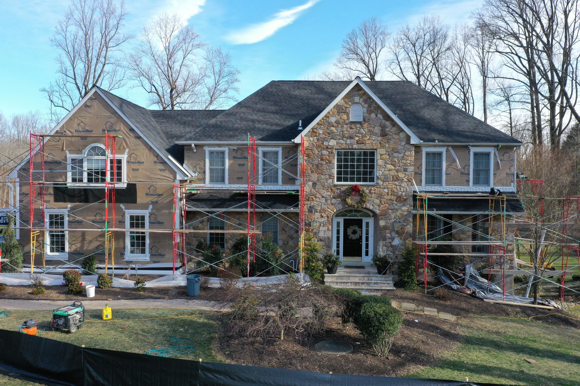 House under construction; brick and stone facade, scaffolding, lawn, trees, blue sky.