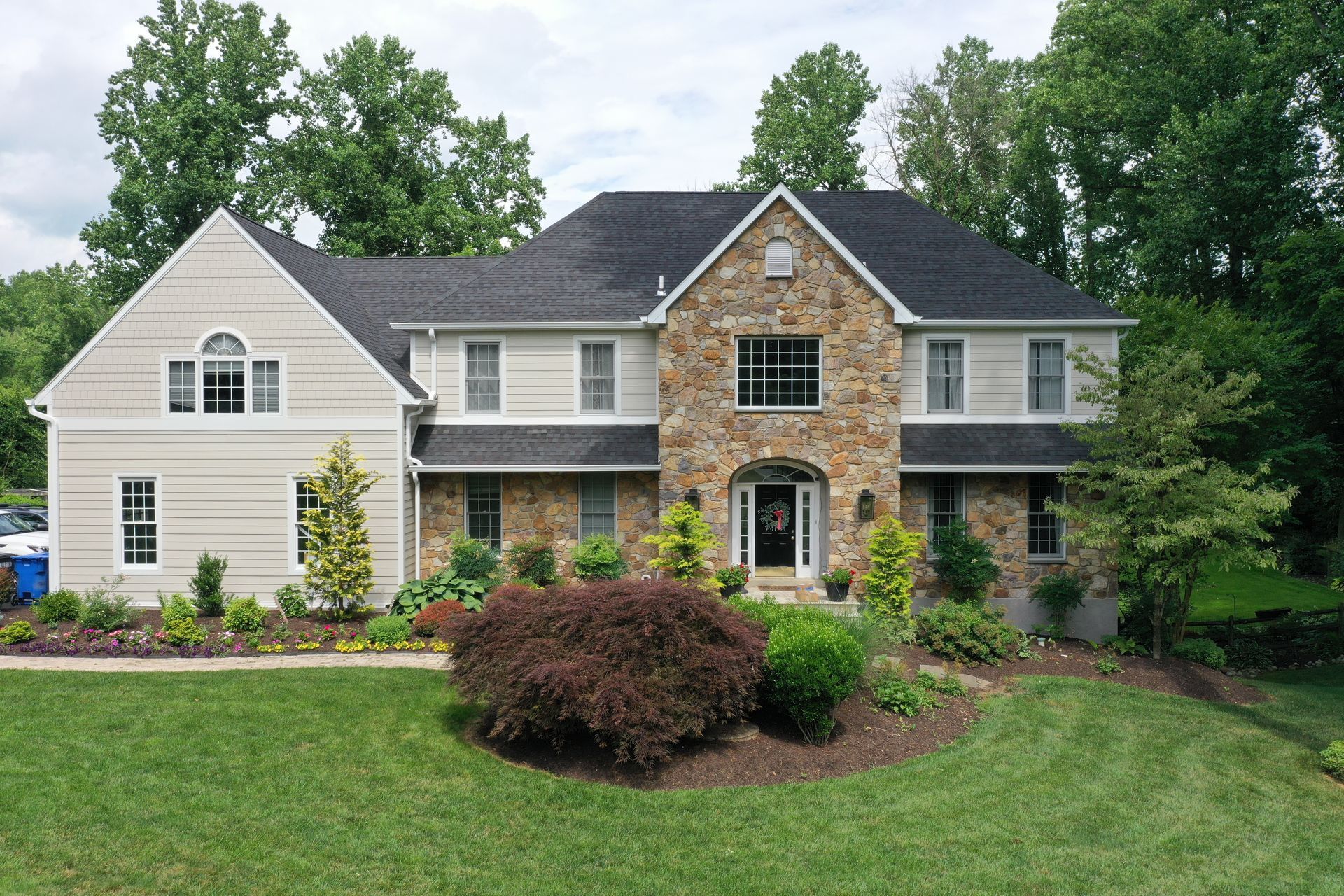 Two-story house with stone facade, beige siding, and black roof, surrounded by green grass and trees.