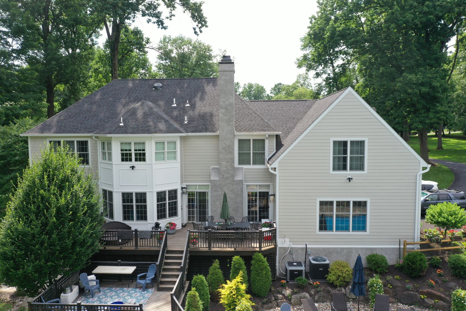 Back view of a light beige house with a chimney and a wooden deck surrounded by trees.