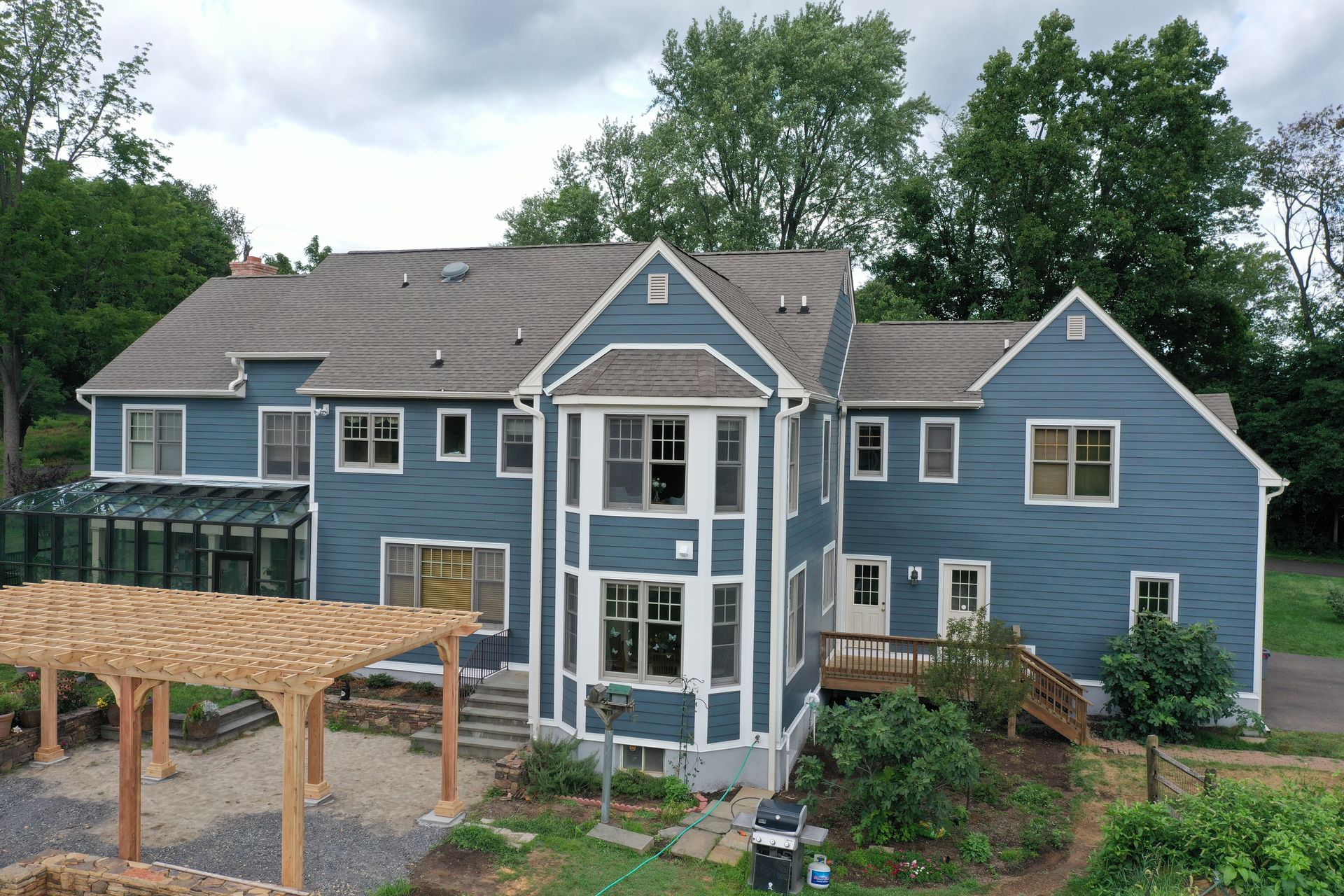 Blue house with multiple stories, windows, and a pergola in front of a wooded area.