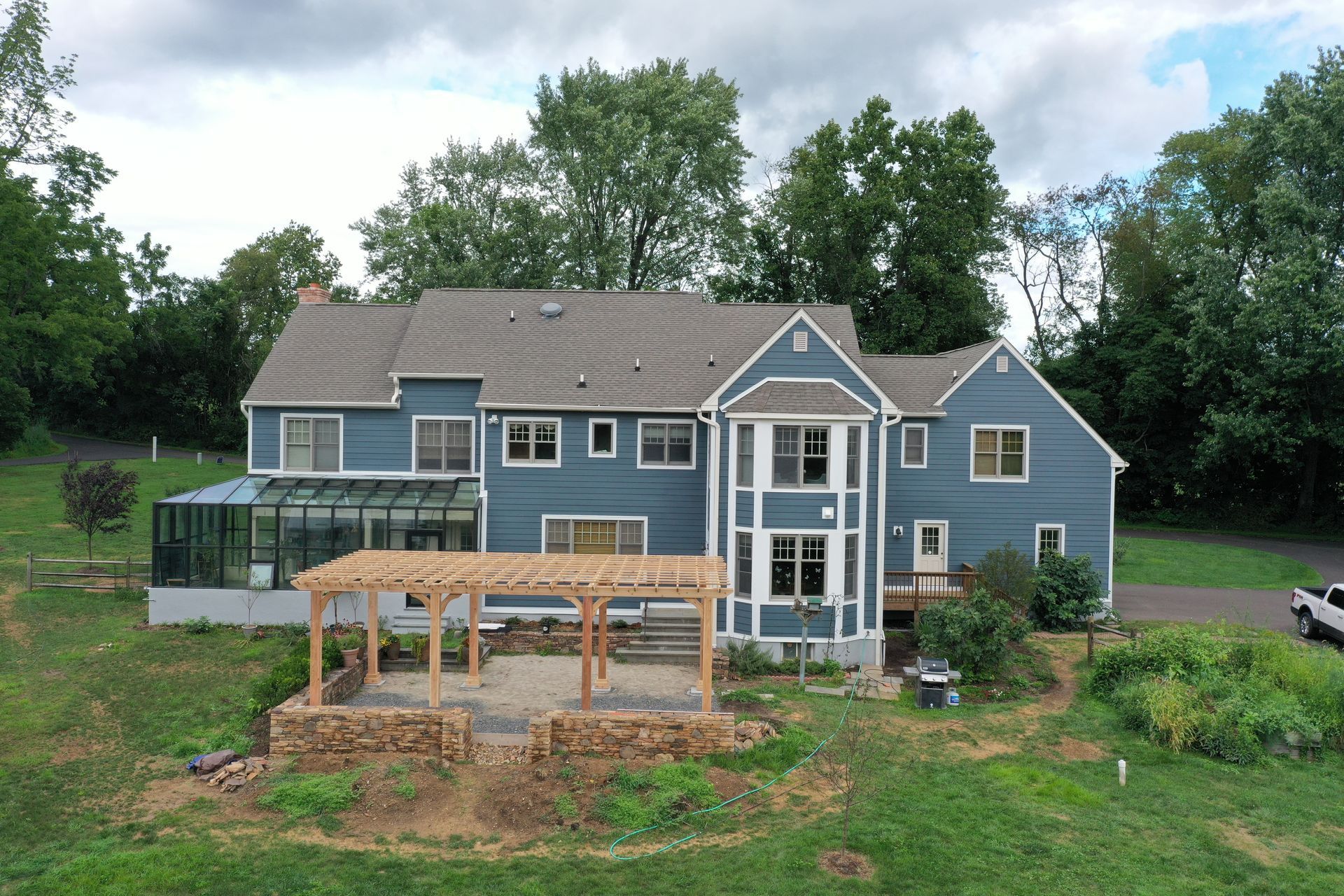 Blue house with pergola, deck, and attached greenhouse, set in a grassy yard, surrounded by trees.