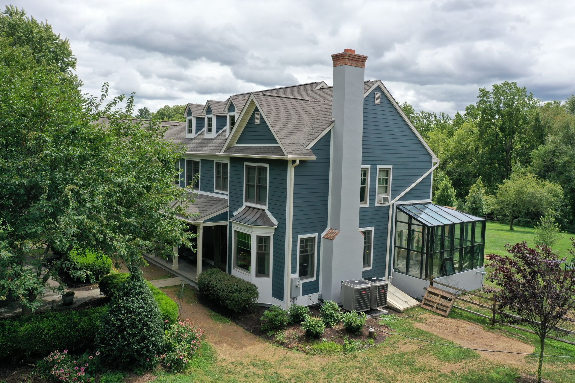 Blue two-story house with a sunroom, chimney, and landscaped yard under a cloudy sky.