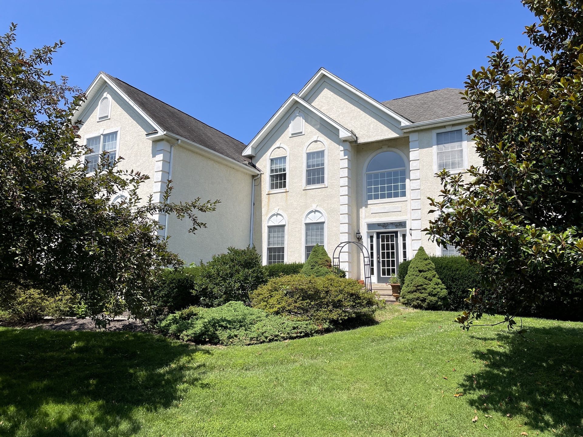 Two-story house with stucco exterior, manicured lawn, and evergreen bushes. Blue sky.