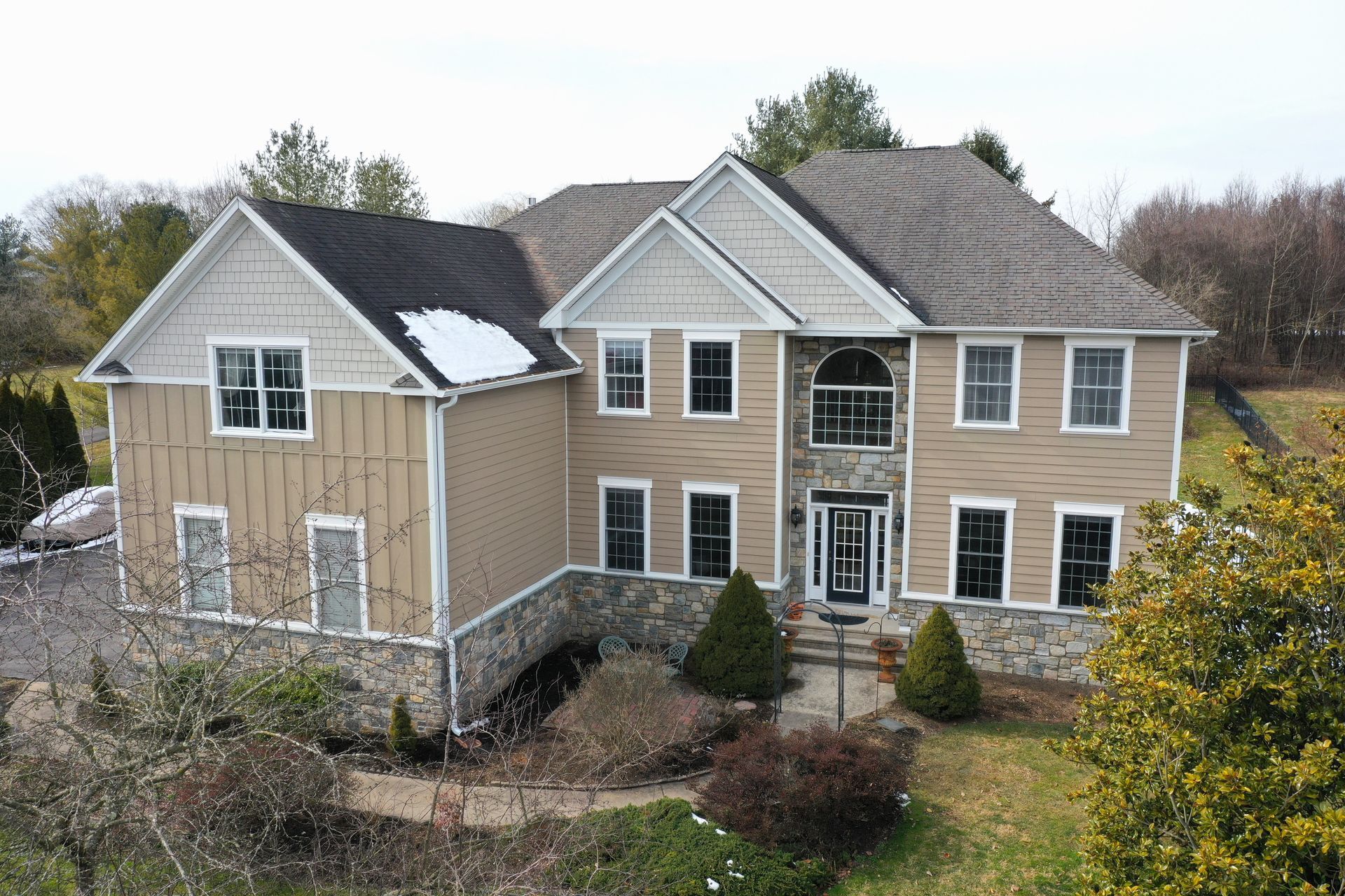 Two-story beige house with stone accents, windows, and a partially snow-covered roof.