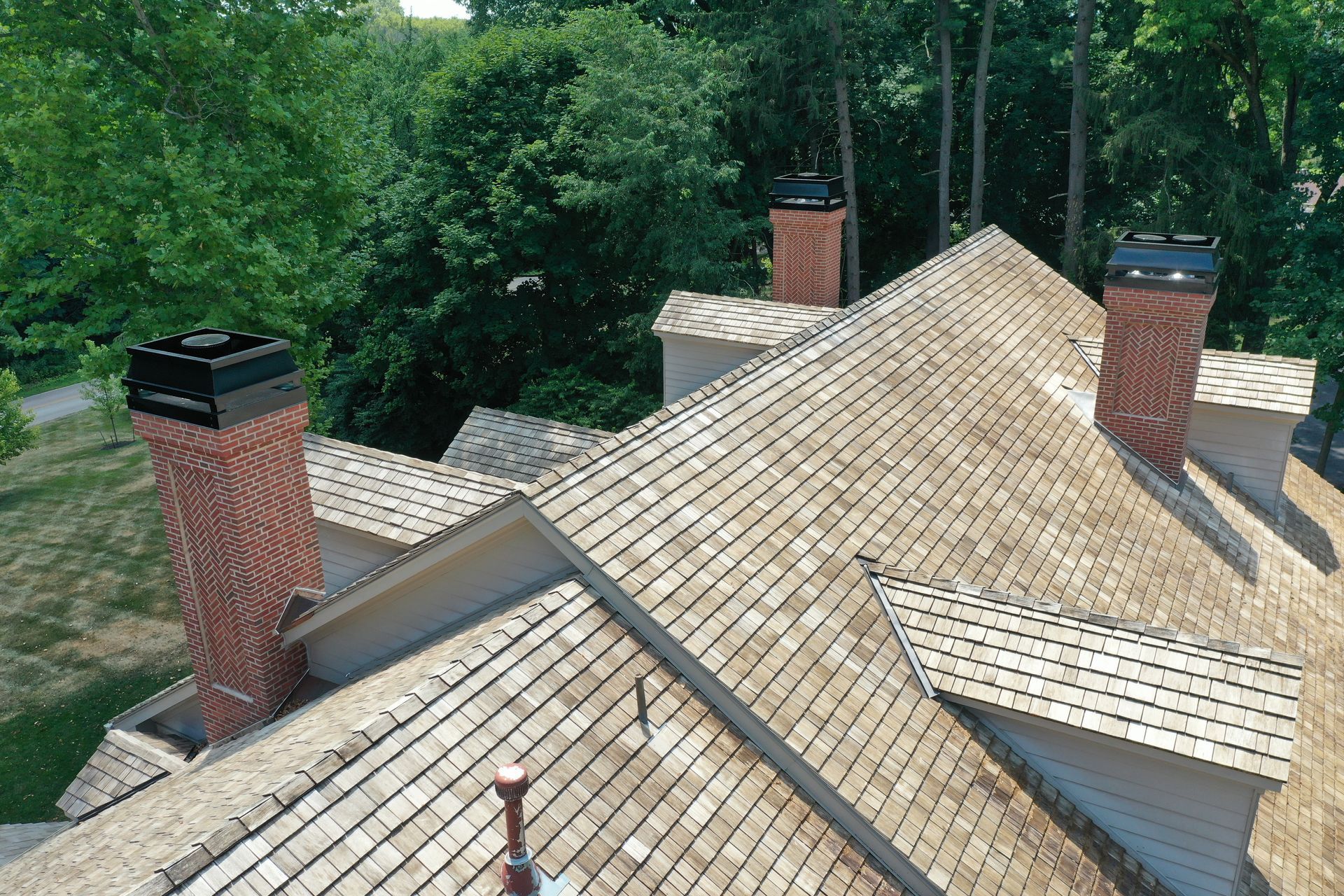 View of a wood shingled roof with three brick chimneys, set against a background of green trees.