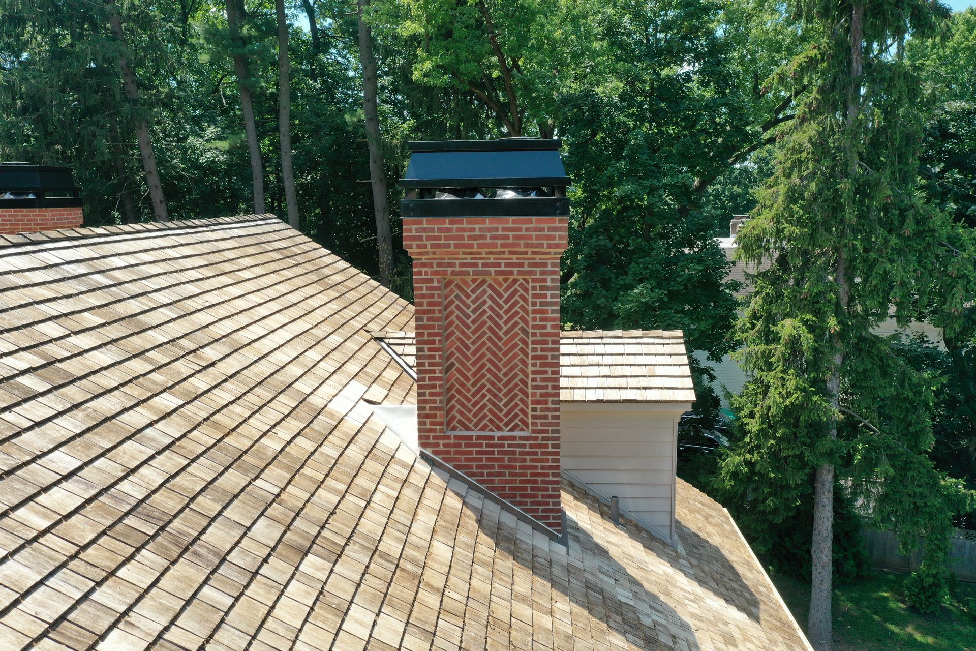 Brick chimney on a shingled roof, trees in the background, topped with a black cap.