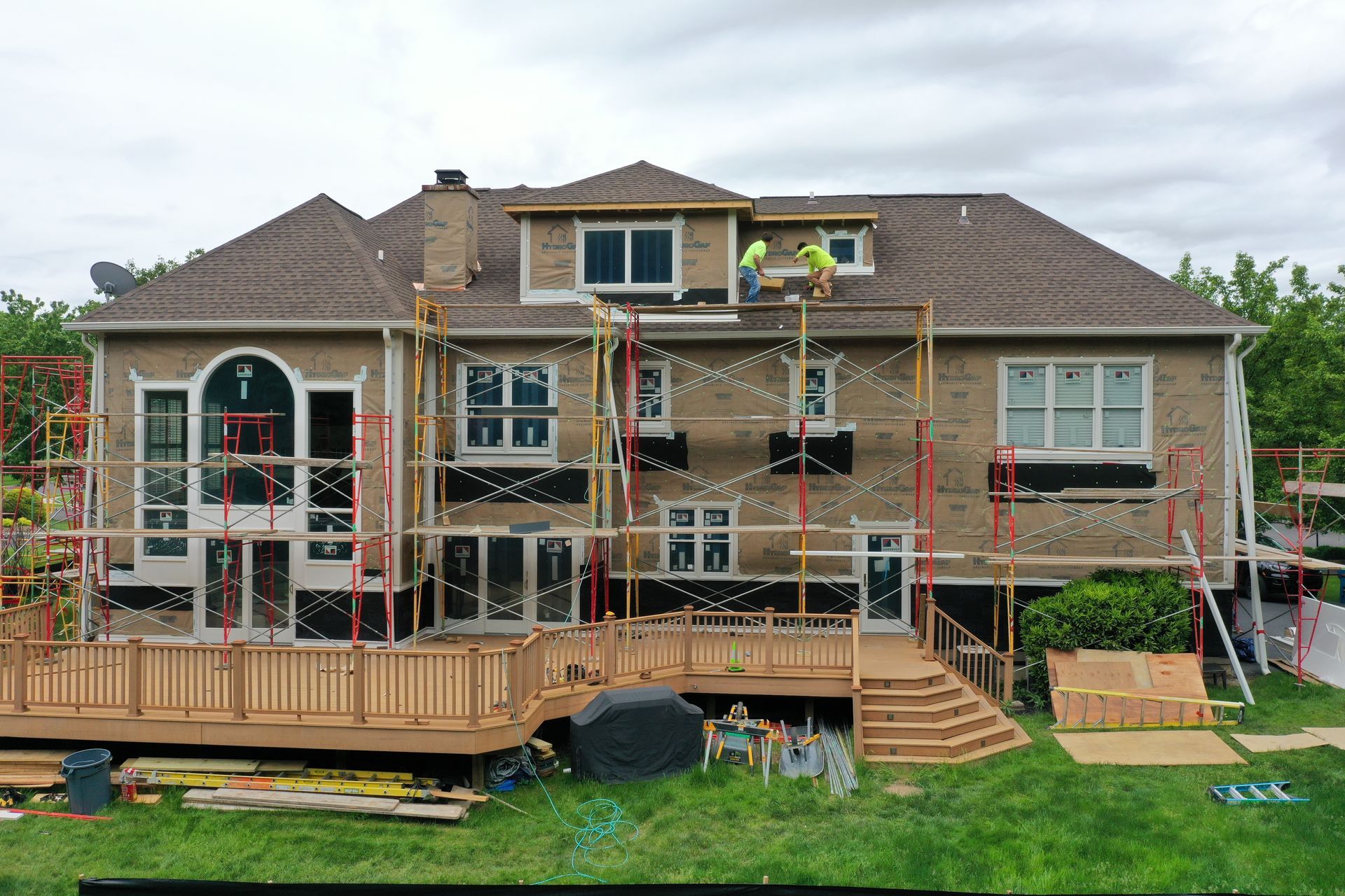 Rear view of a house under construction with scaffolding and workers on the roof. Wooden deck and green lawn visible.