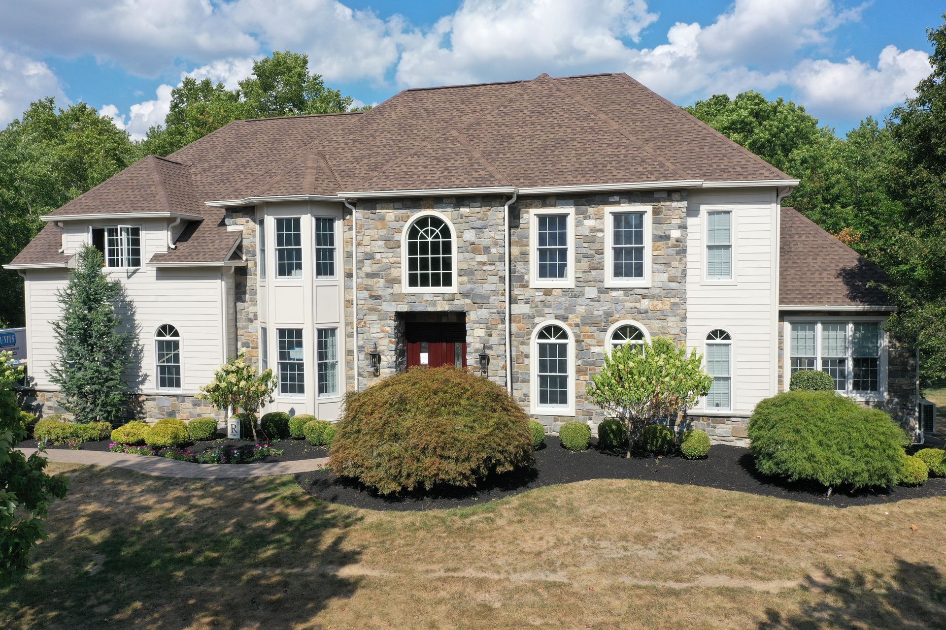 Large two-story house with stone facade, light siding, brown roof, and landscaping under a blue sky.