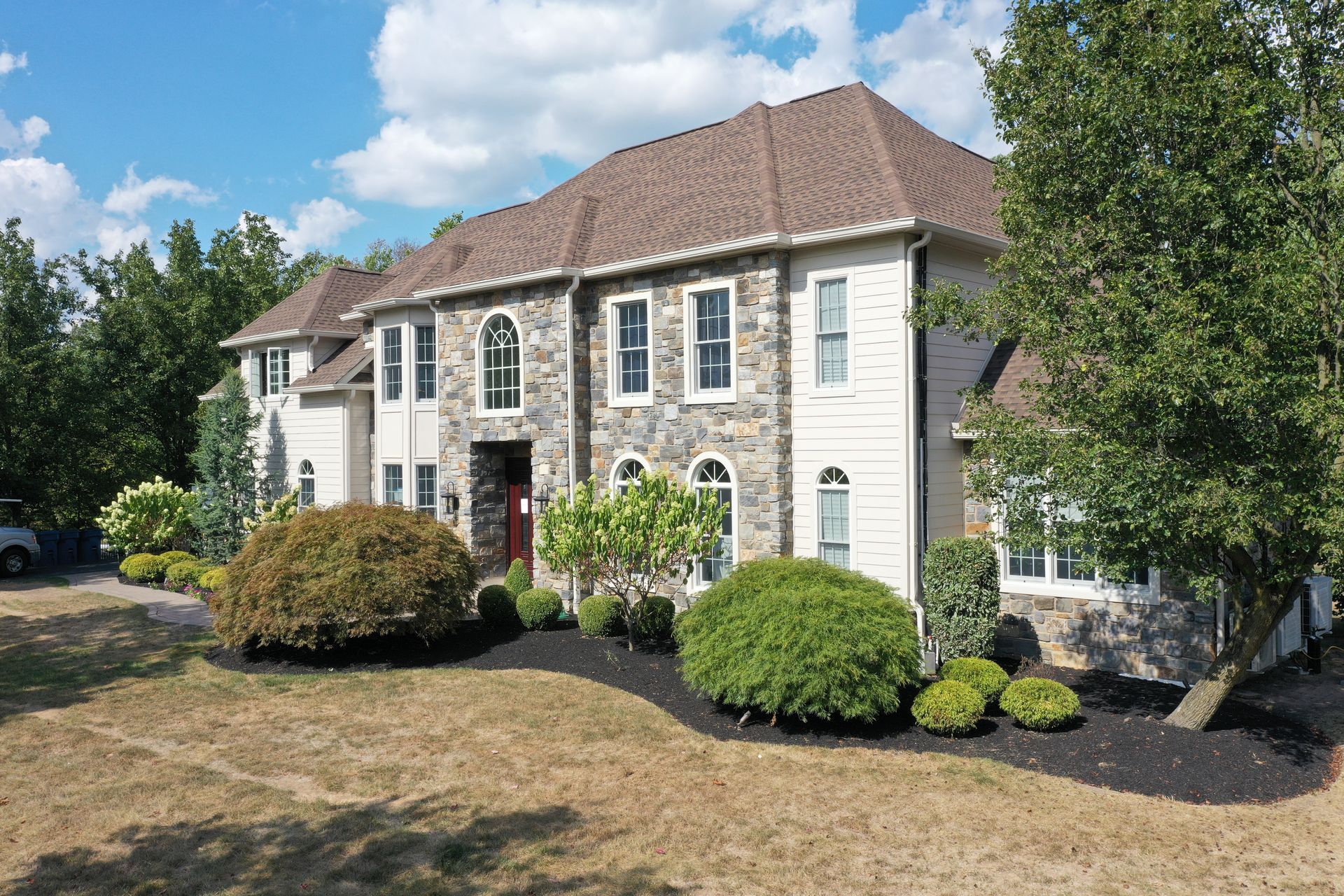 Large two-story house with stone facade, tan siding, and brown roof under a blue sky, surrounded by landscaping.