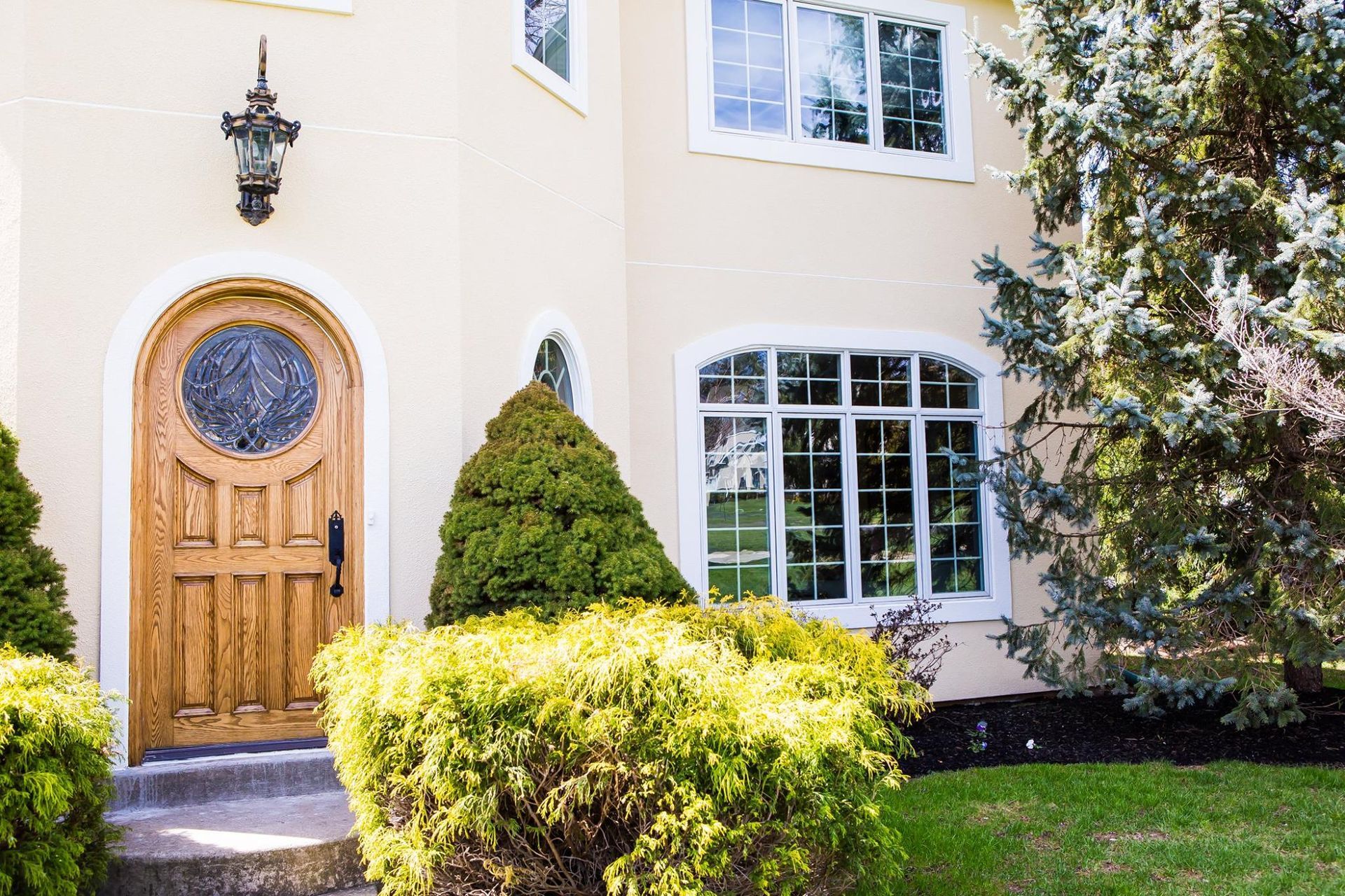 Beige stucco house with arched wooden door, multi-pane windows, and manicured landscaping.