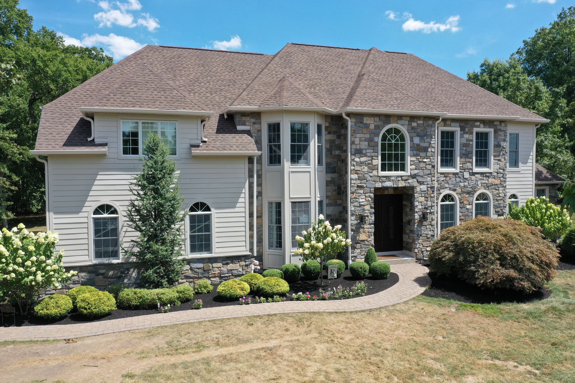 Two-story house with stone and siding facade, curved walkway, and landscaping under a blue sky.