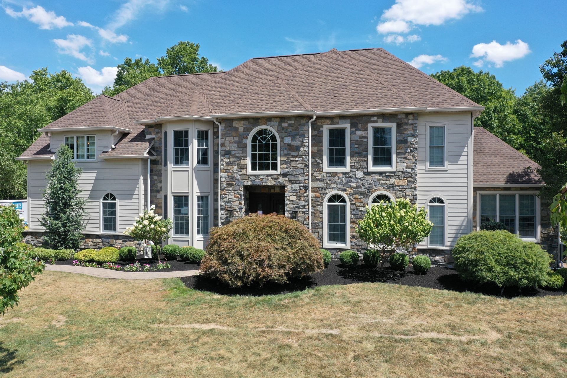 Large two-story house with stone facade, light siding, brown roof, and arched windows, surrounded by trees and lawn.