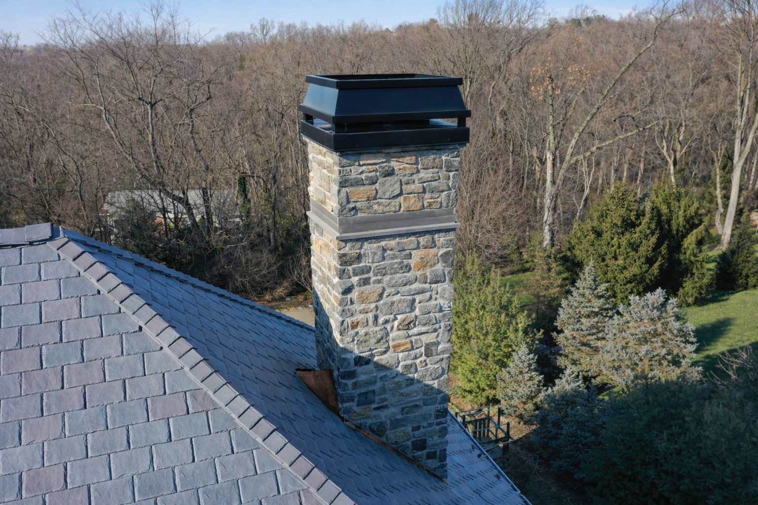 Stone chimney on a slate roof, topped with a black cap, set against bare trees and green shrubs.