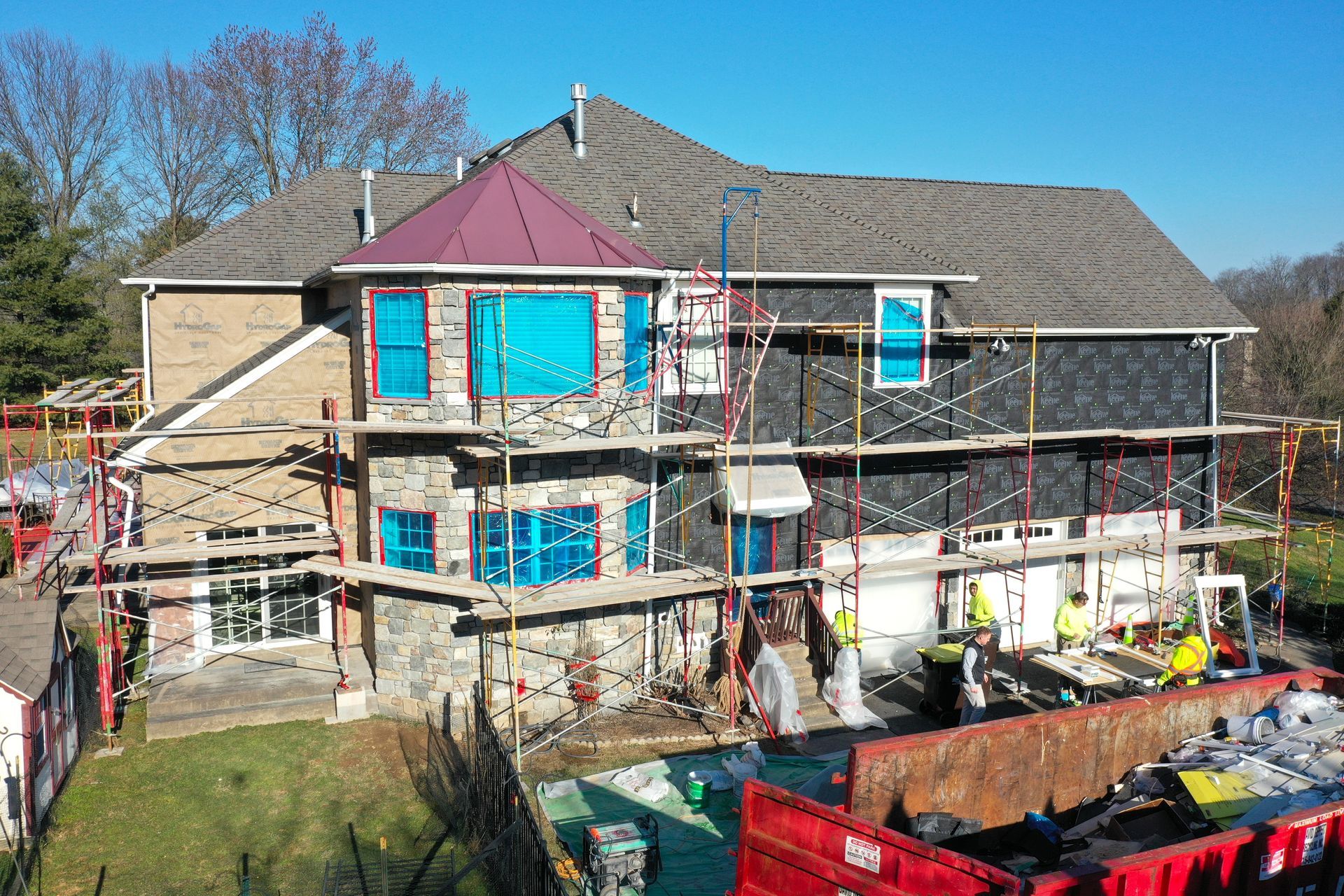 House under construction, scaffolding surrounding the structure. Workers in yellow vests near a red dumpster.