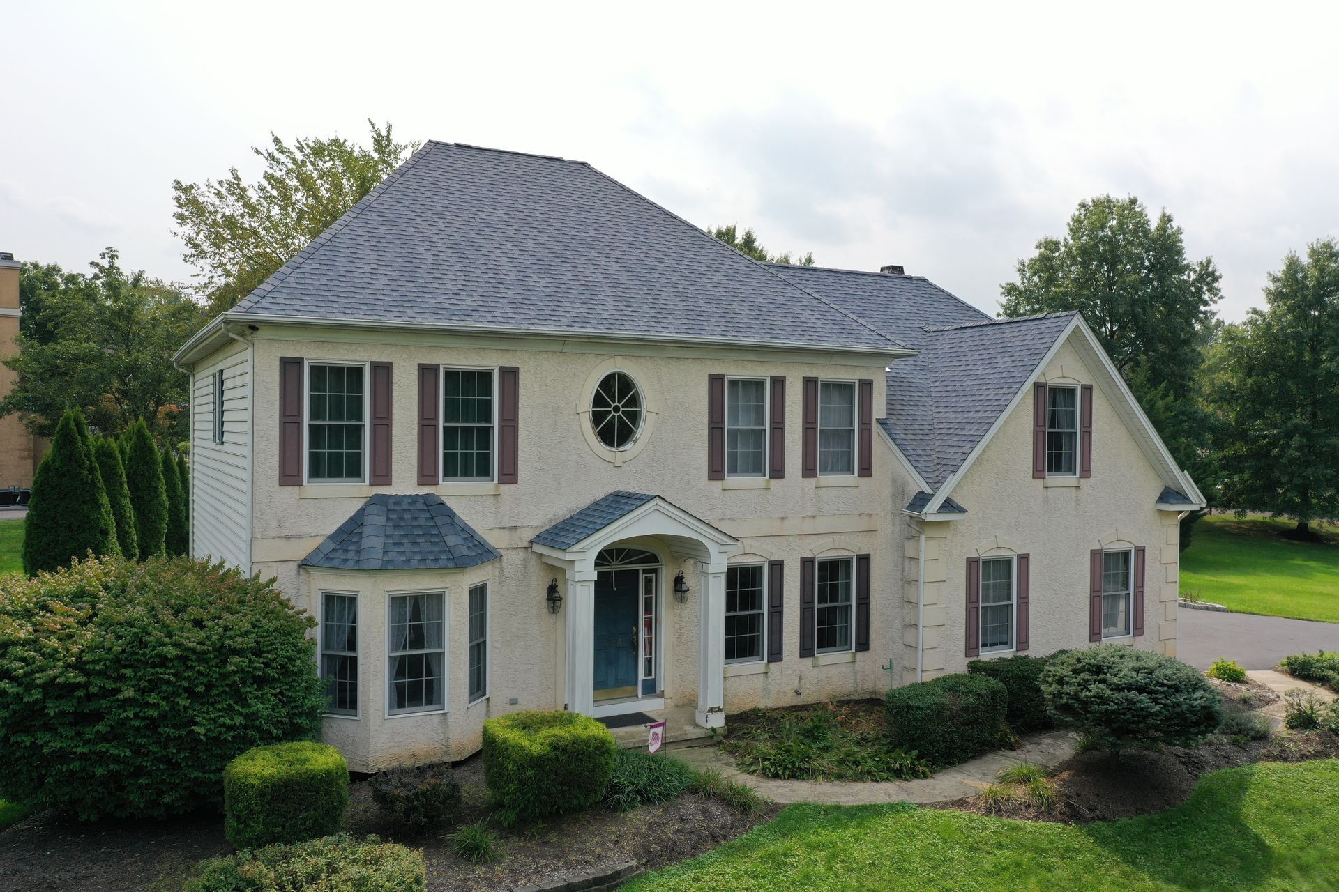 Two-story beige house with gray roof, brown shutters, and green bushes.