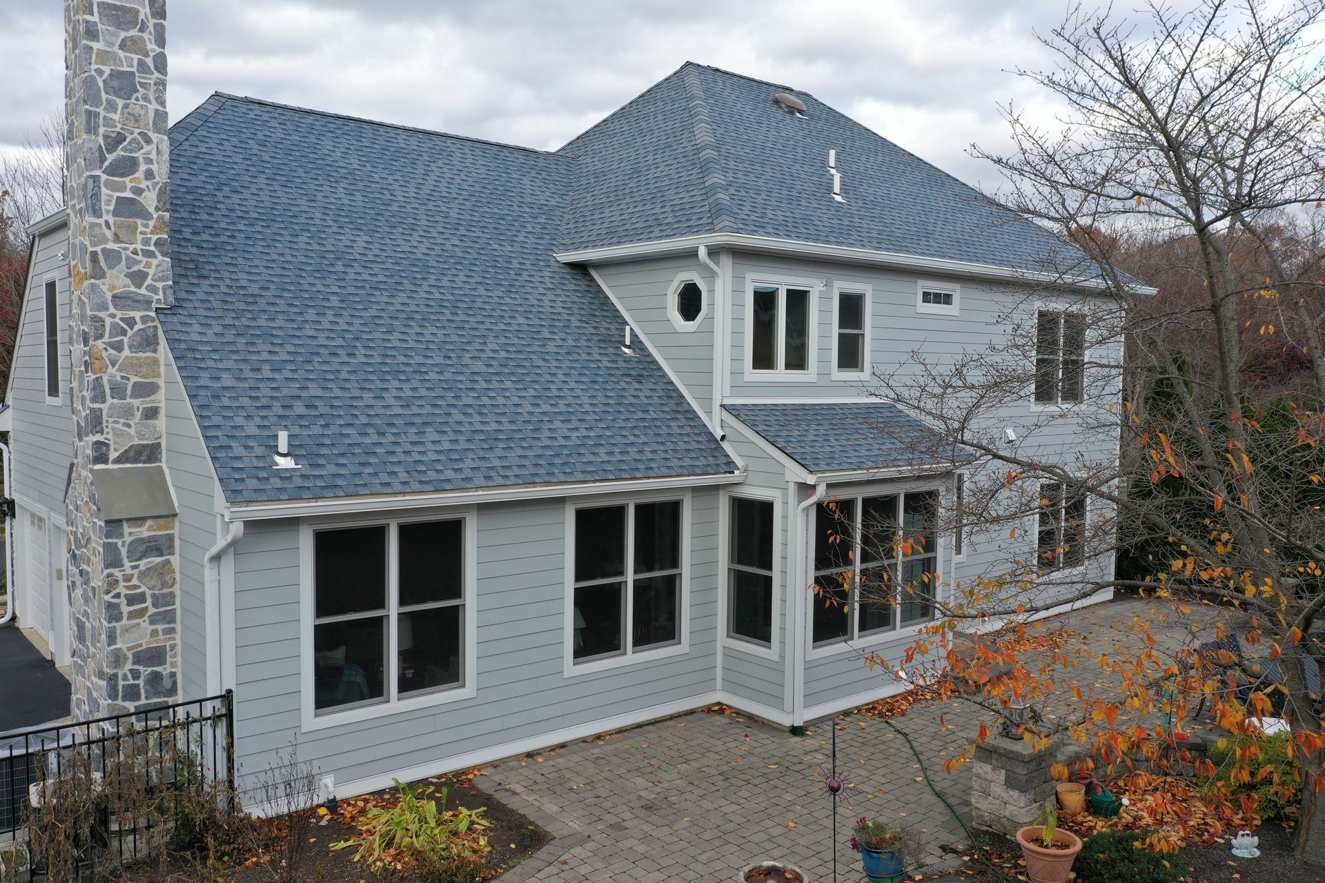 Two-story house with gray siding, blue roof, stone chimney, and large windows on a paved patio.