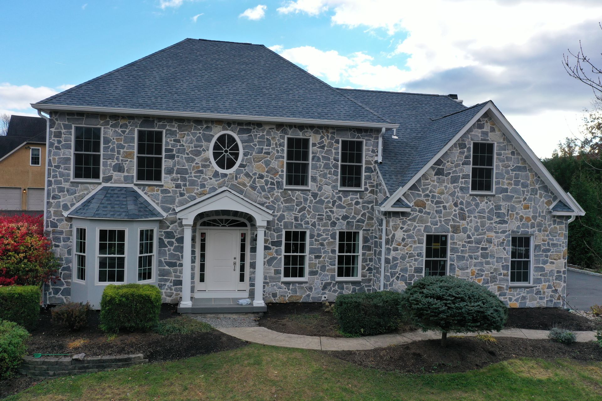 Two-story stone house with a gray roof and numerous windows, set on a grassy lawn with manicured bushes.
