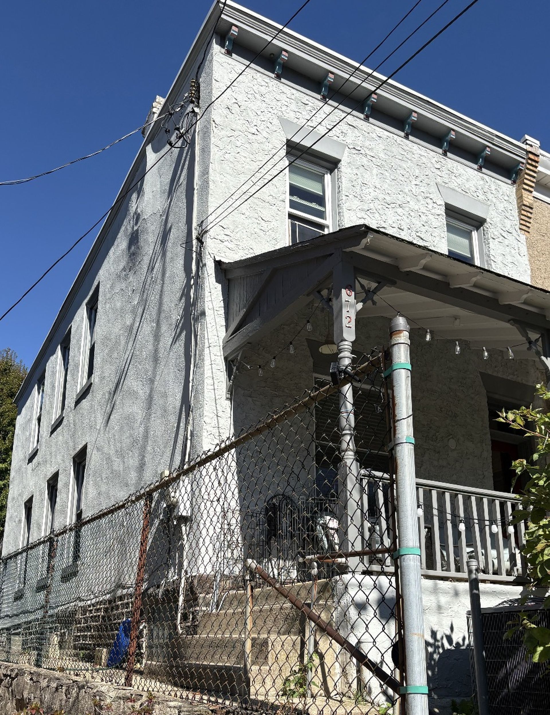Two-story gray building with a porch, a chain-link fence, and stairs leading up. Clear blue sky.