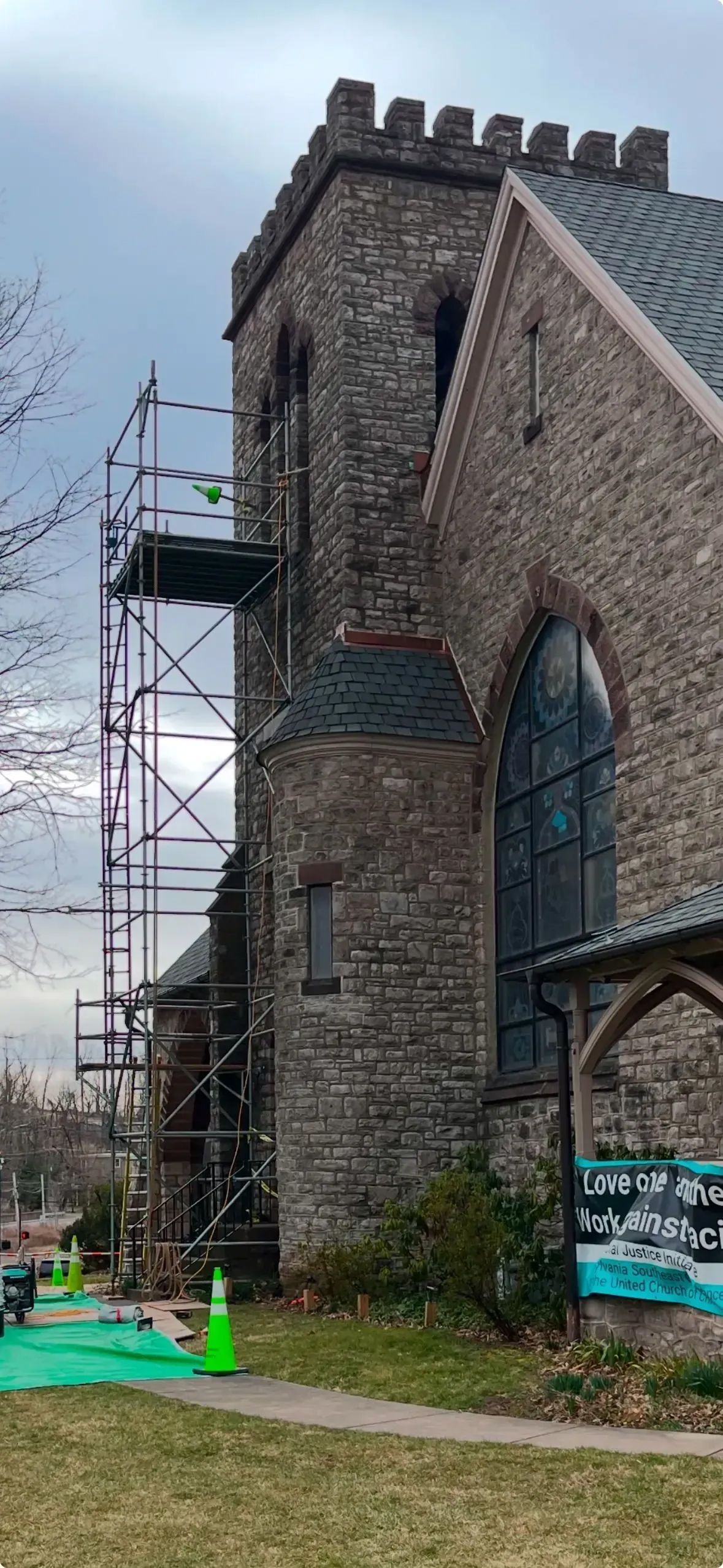 A stone church with a tall tower has scaffolding set up for work; green turf and cones in front.