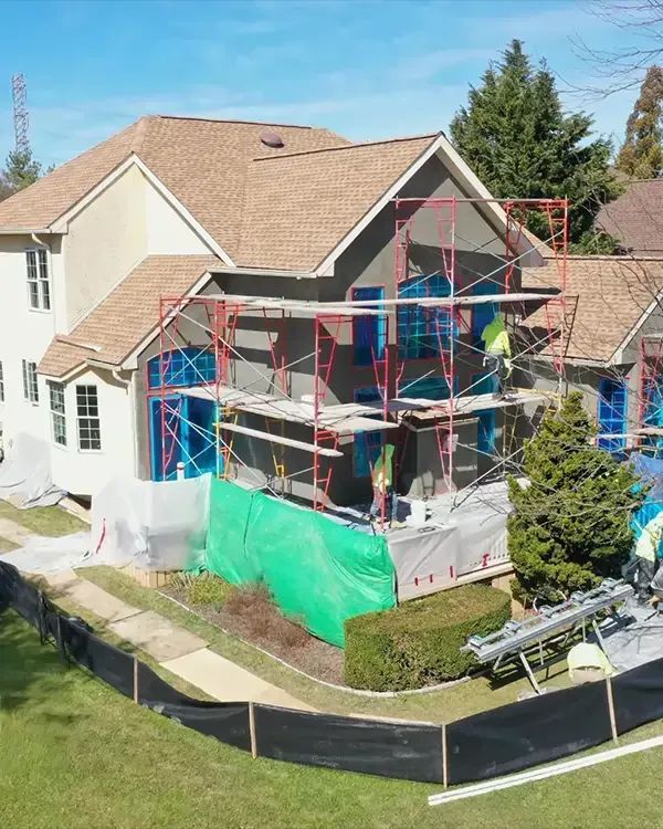 House exterior under construction, scaffolding, blue tarp-covered windows, green protective sheeting, brown roof.