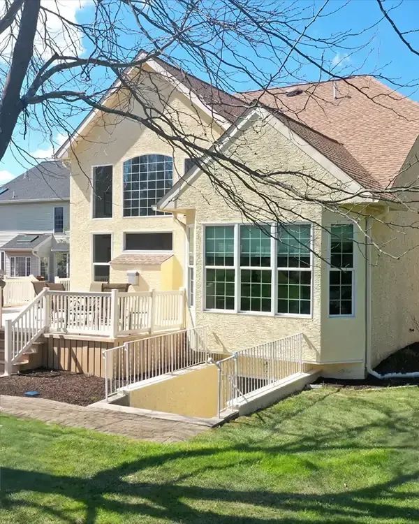 Beige house with deck, large windows, and a basement entrance, set on a green lawn.