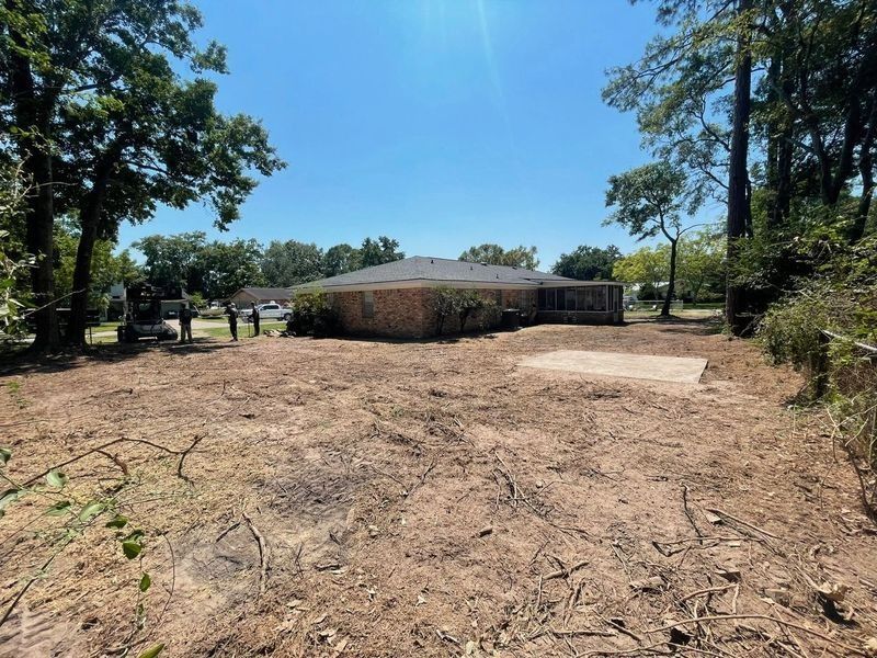A cleared lot with a brick house in the background, trees frame the sky, and sunlight shines.