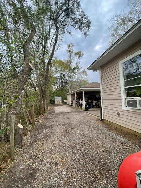 Gravel driveway leading to a house with beige siding and a covered parking area; trees and overcast sky.