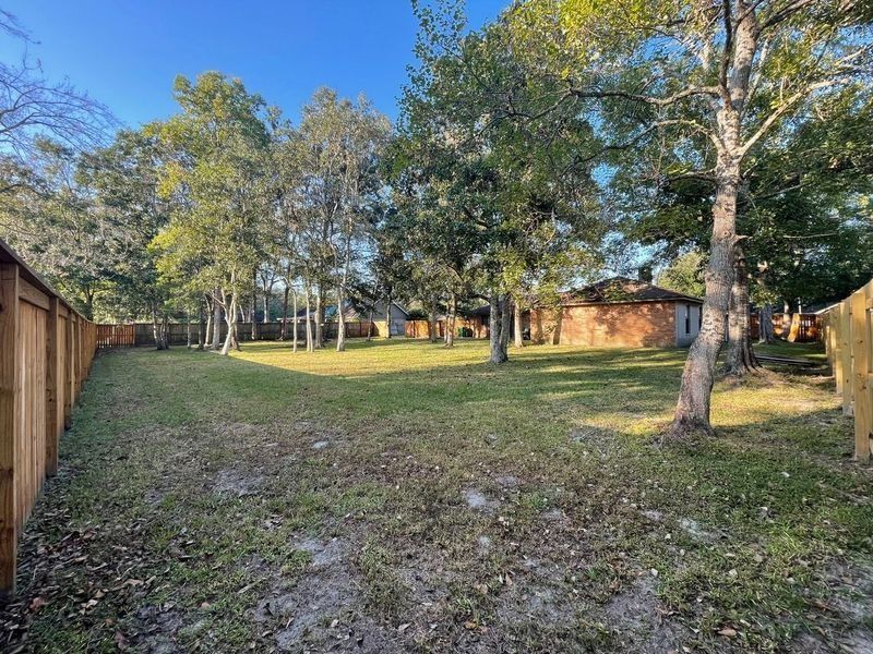 Grassy backyard with brown fence on either side, trees in the middle, and a red brick house in the background.