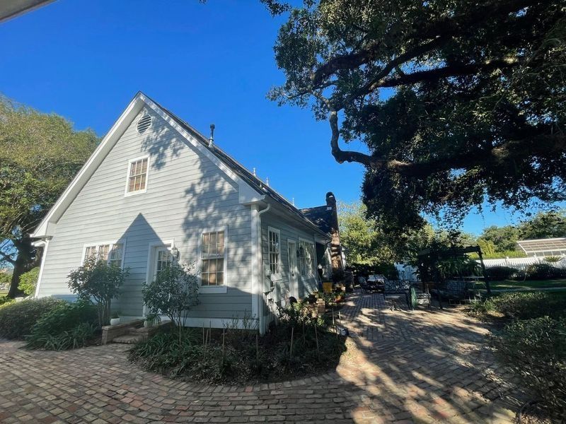 A gray house with a brick walkway under a bright blue sky, partially shaded by a tree.
