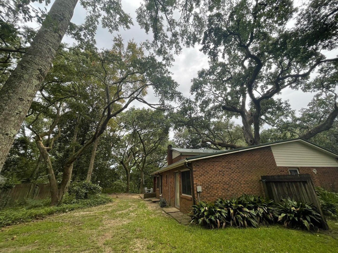 Brick house nestled among large trees on a cloudy day. Green grass and foliage in the foreground.