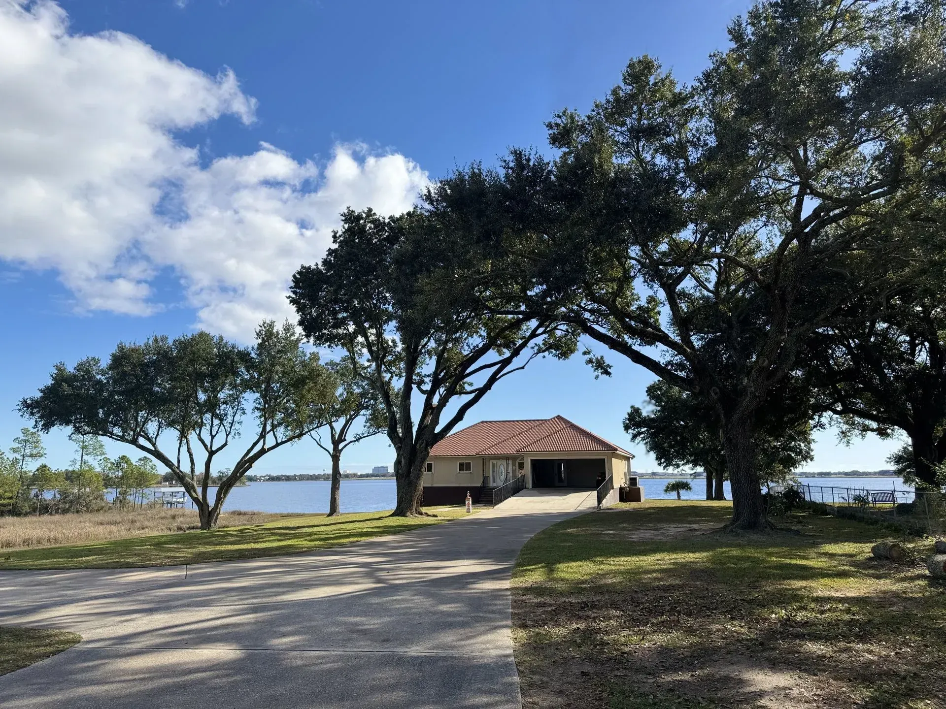 A house with a brown roof sits on a body of water with trees and a driveway. Blue sky with clouds.