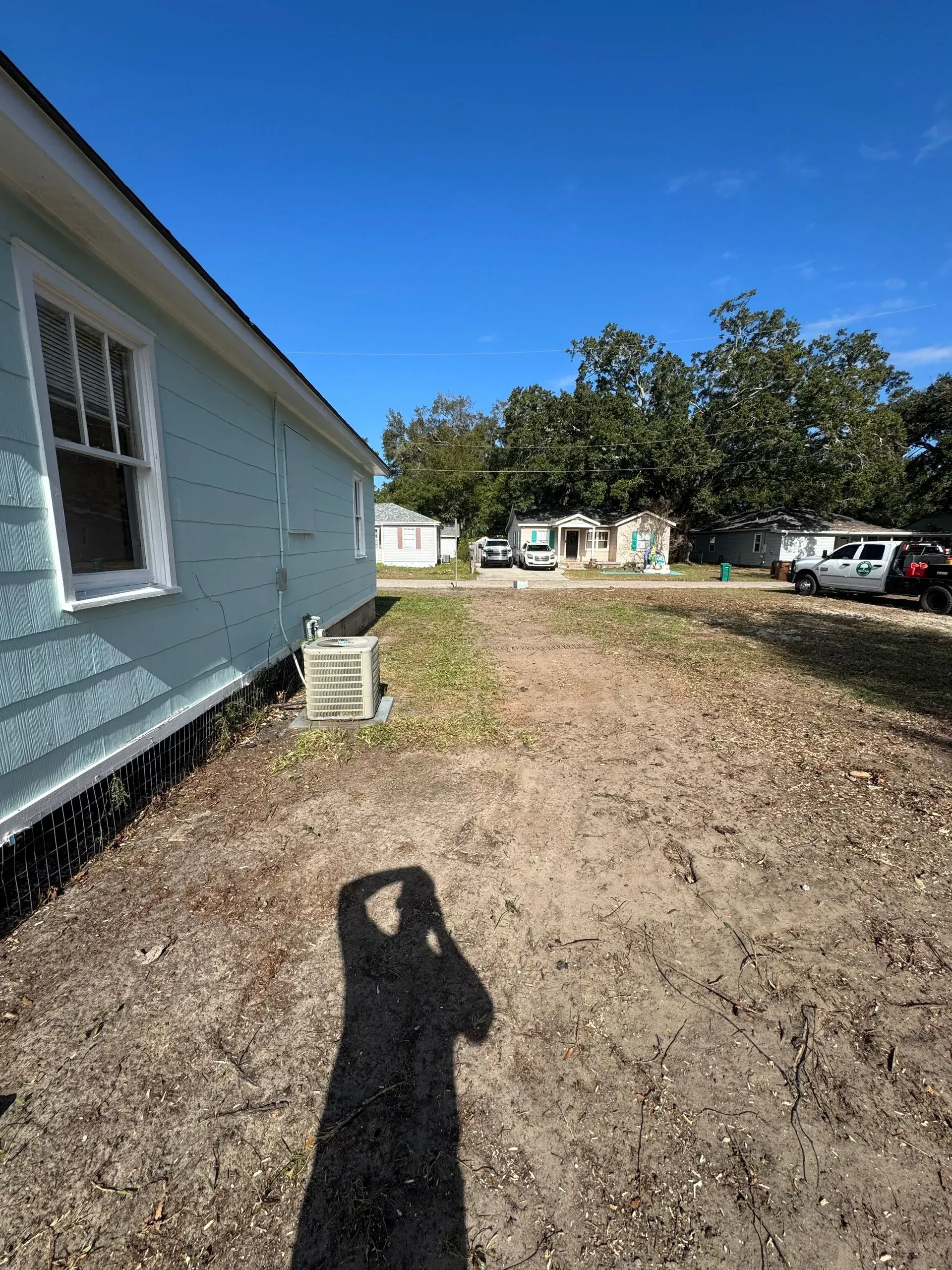 Side view of a light blue building with a window, dry brown ground, and other houses in the distance under a blue sky.