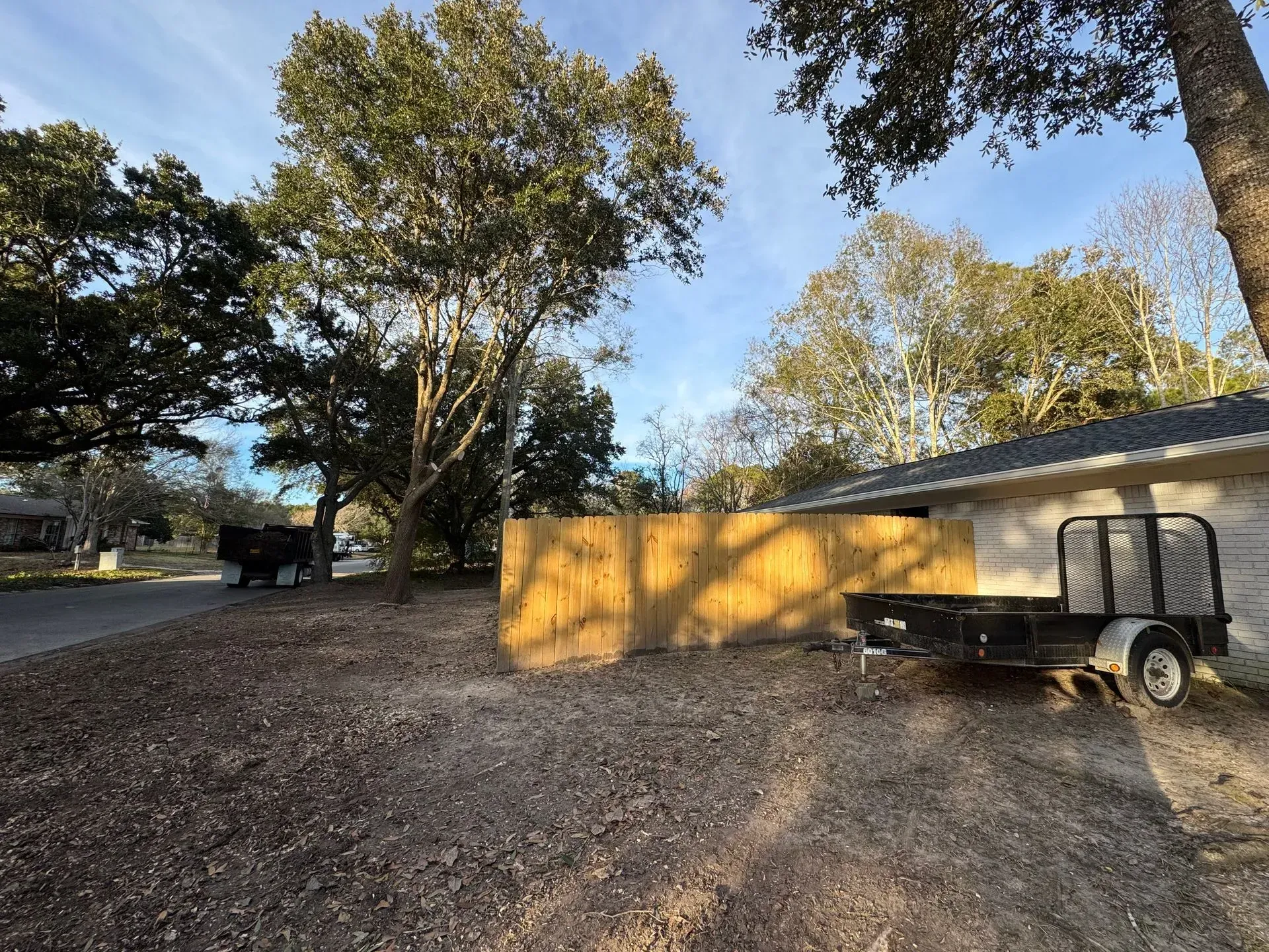 A dirt yard with a wooden fence, trailer, and trees against a house and blue sky.