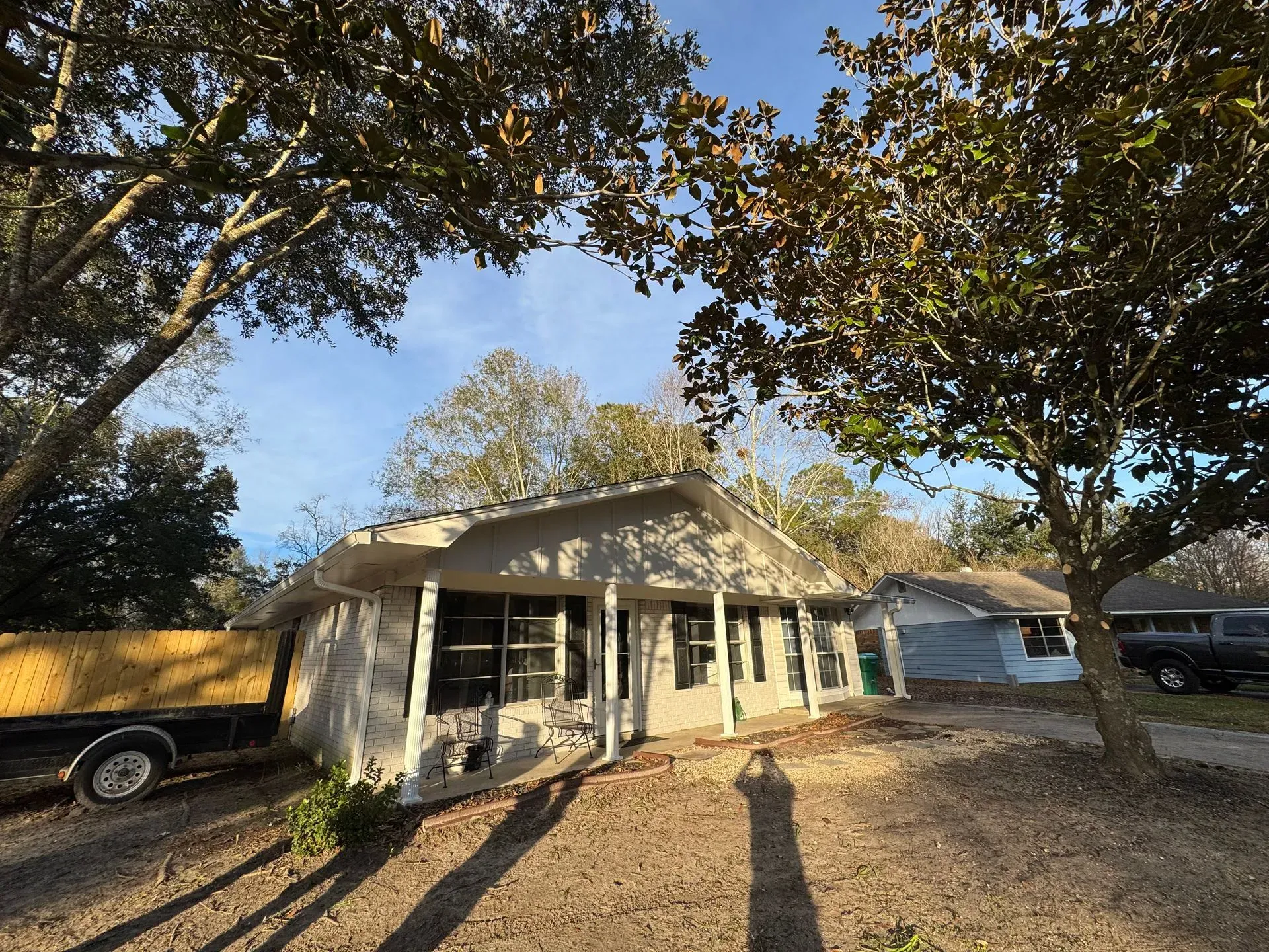 A single-story house with a light-colored exterior and a dark roof, under trees. Sunlight casts long shadows.