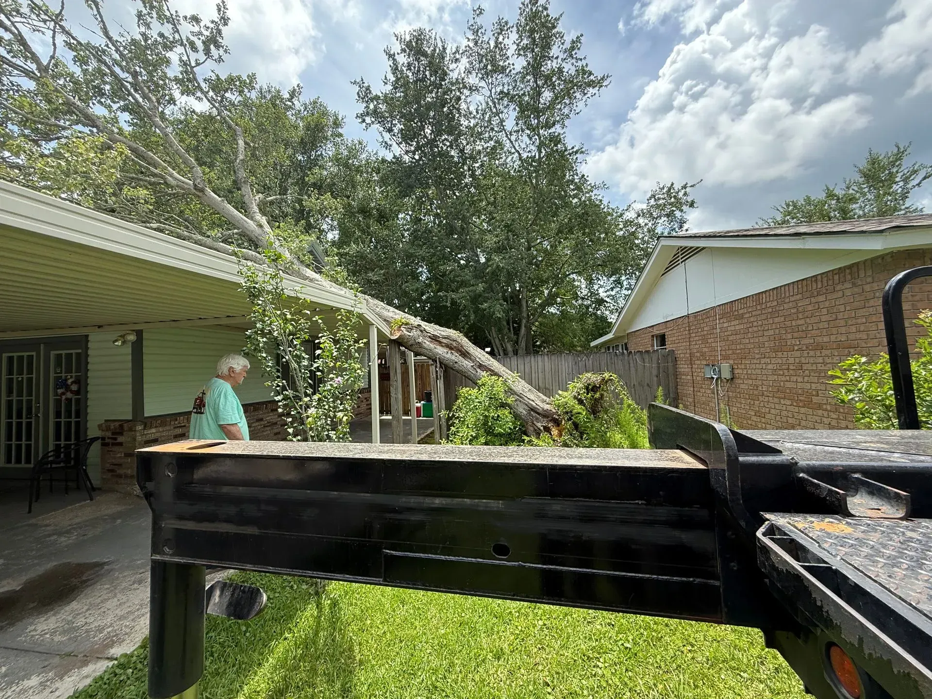 A tree limb rests on a truck bed, next to a home with a person observing. Green grass and trees are in the background.