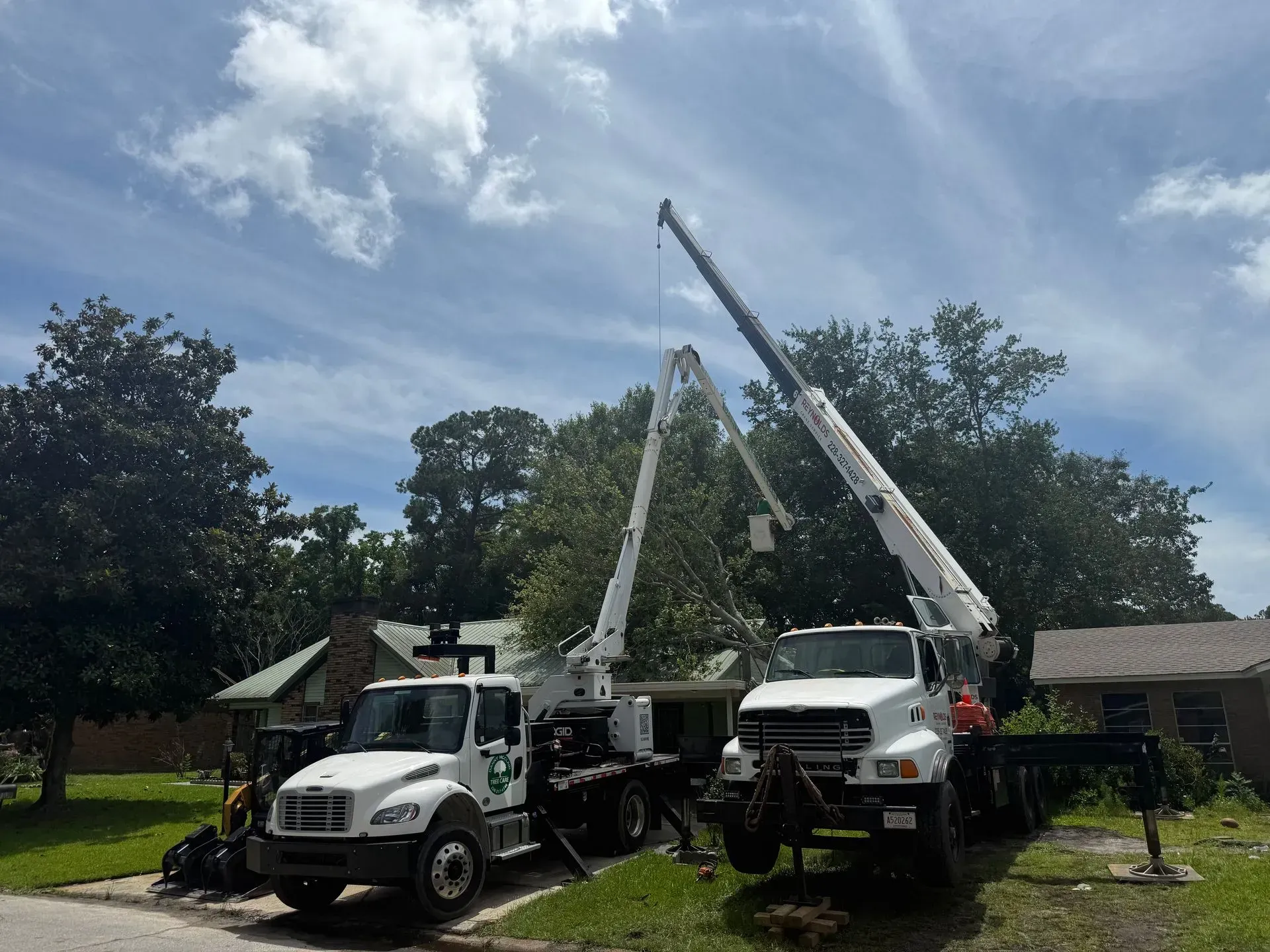 Two tree service trucks with extended booms trimming a tree in front of a house on a cloudy day.