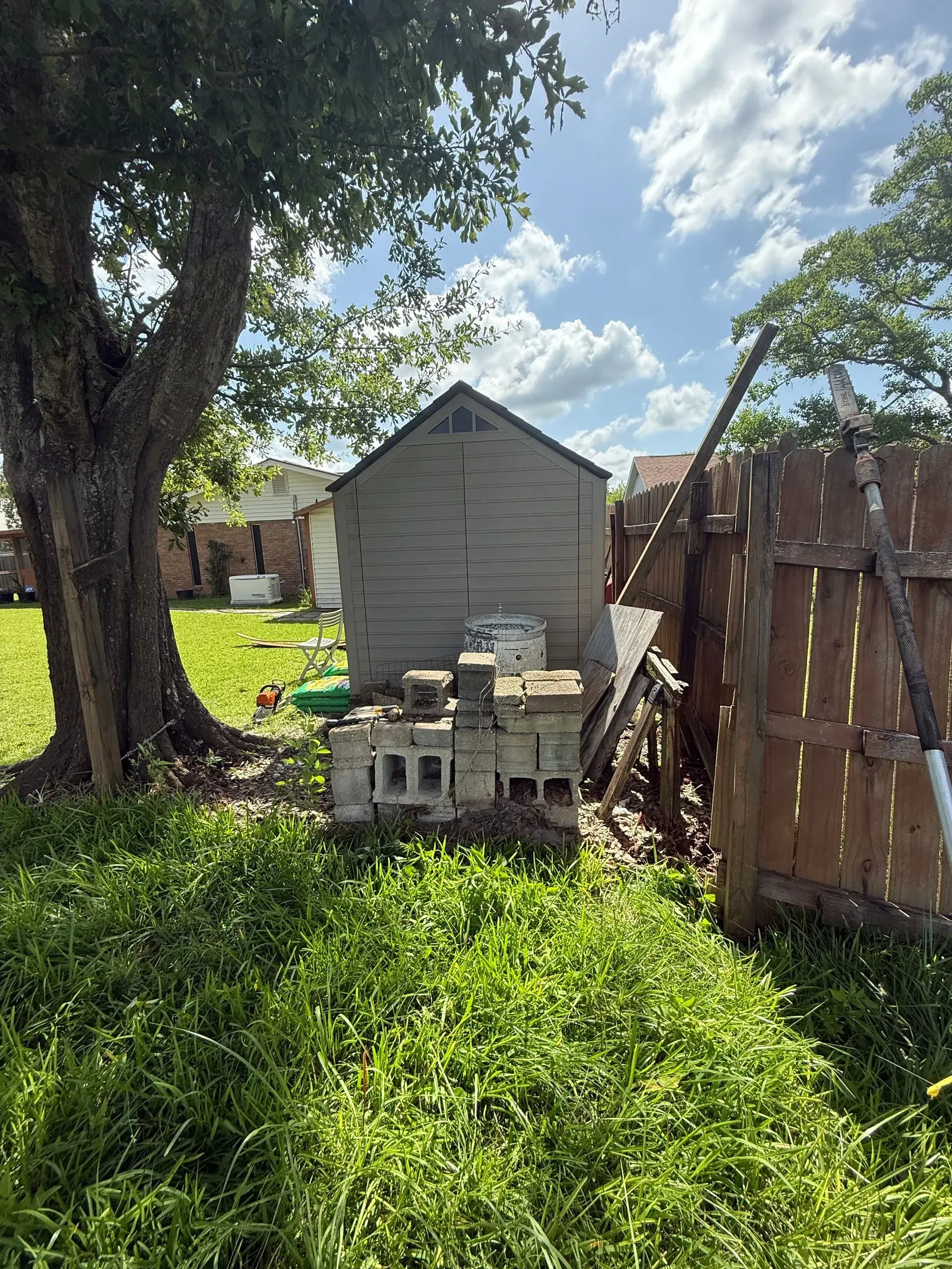 A shed, cinder blocks, and a wooden fence sit in a grassy yard under a tree on a sunny day.