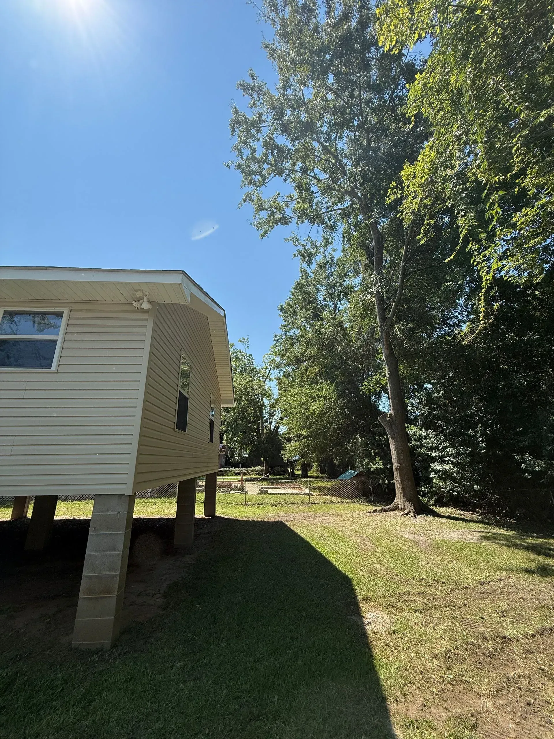 Side view of a raised beige home with trees in a grassy yard under a blue sky.