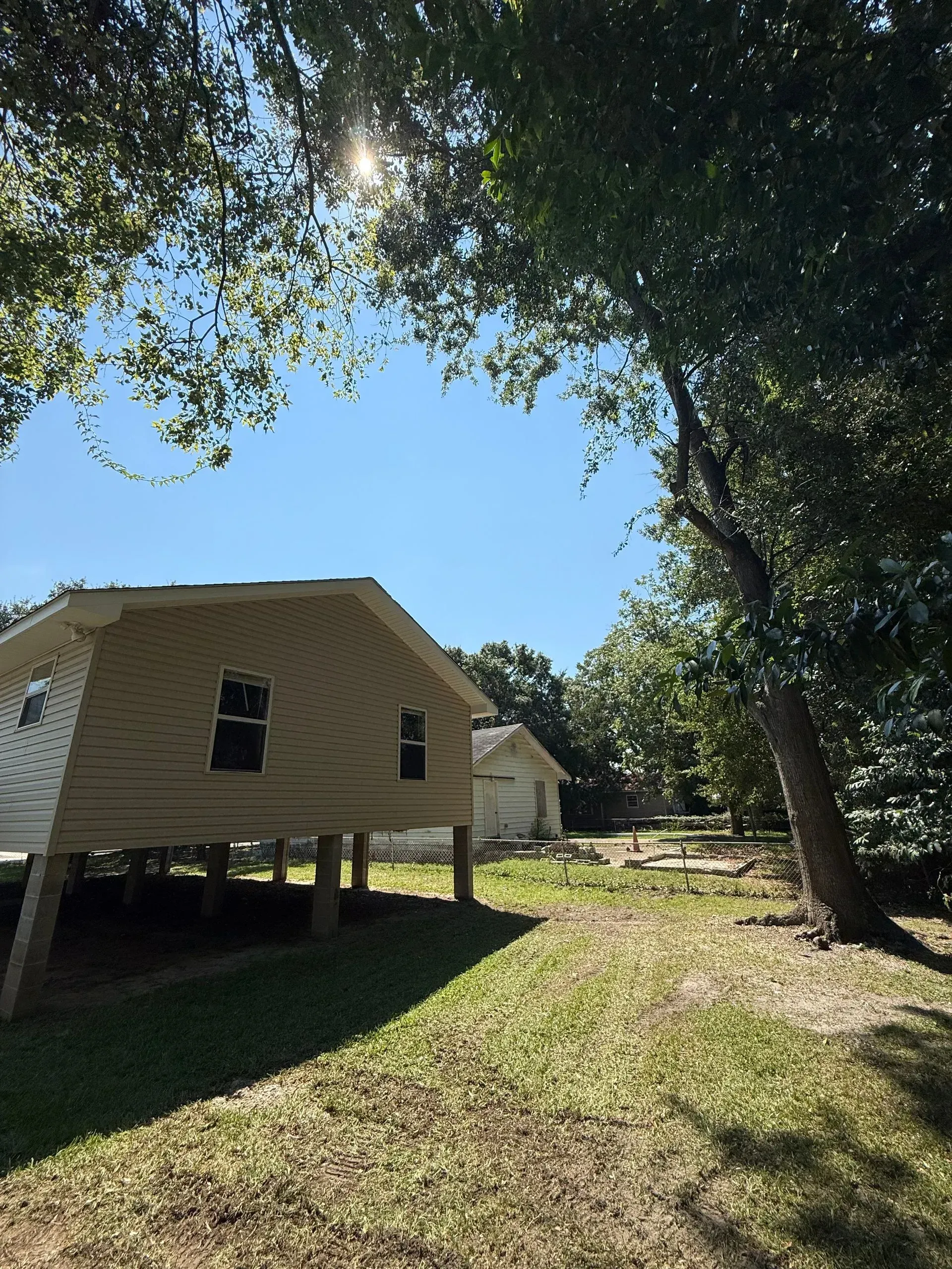 Tan building on stilts, surrounded by trees and grass under a bright blue sky.