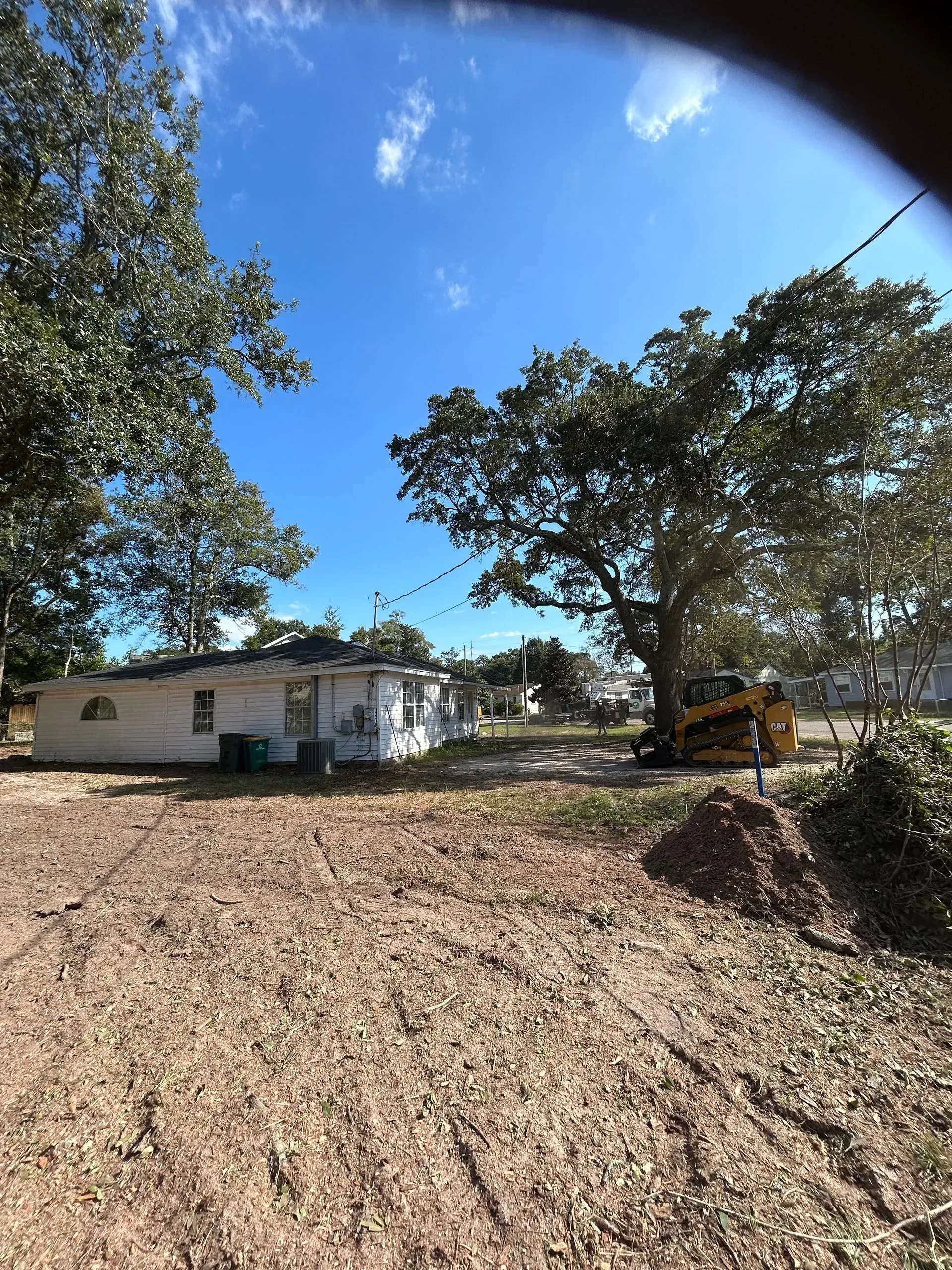 Small white house with landscaping work in progress; trees, blue sky, and a yellow construction vehicle are visible.