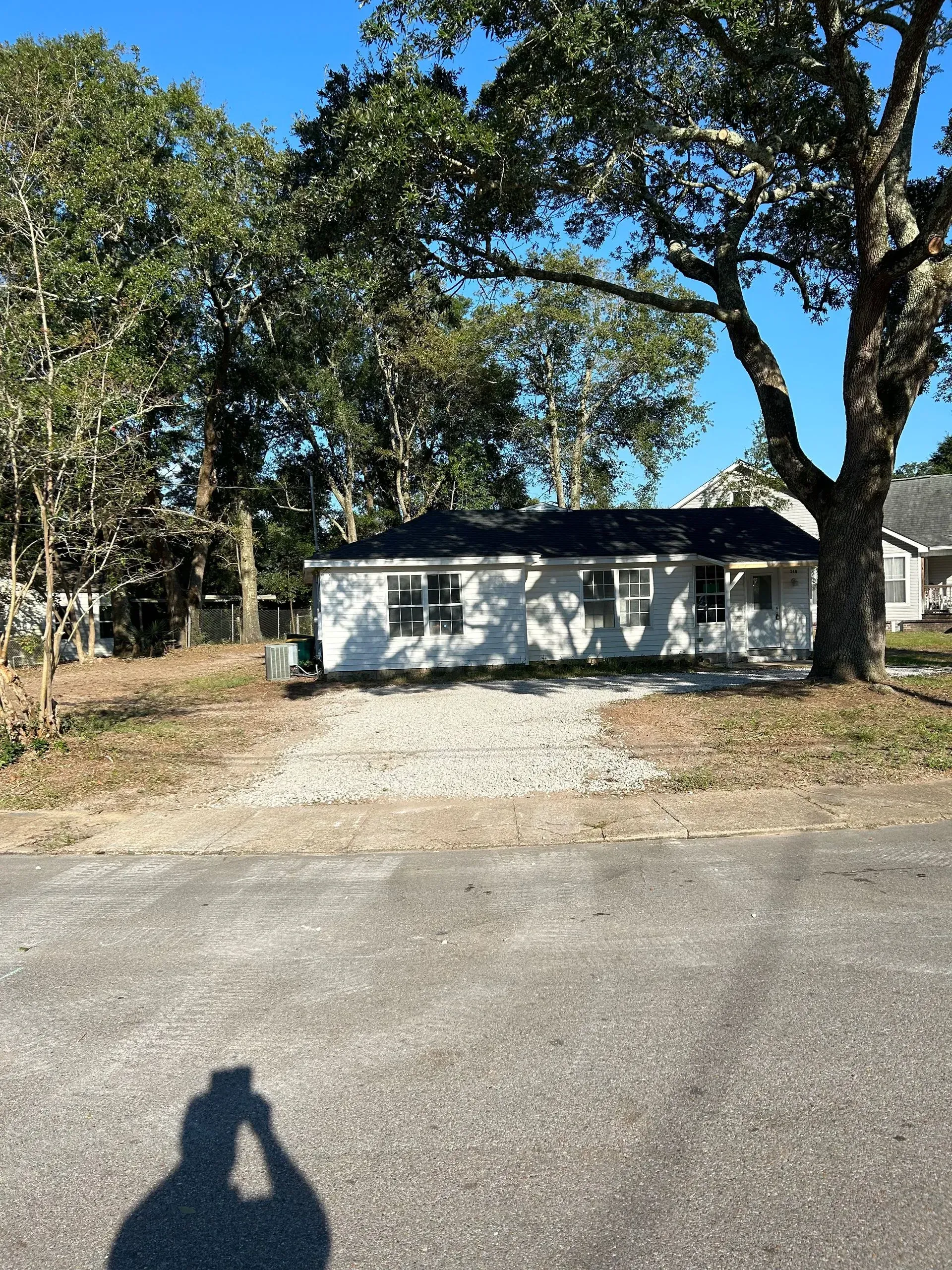 Small, weathered, white house with dark roof, surrounded by trees and a gravel driveway on a sunny day.