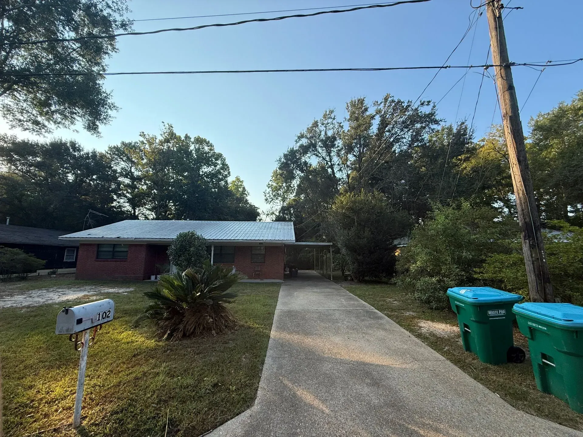 A single-story brick house with a driveway, greenery, and two green trash cans.