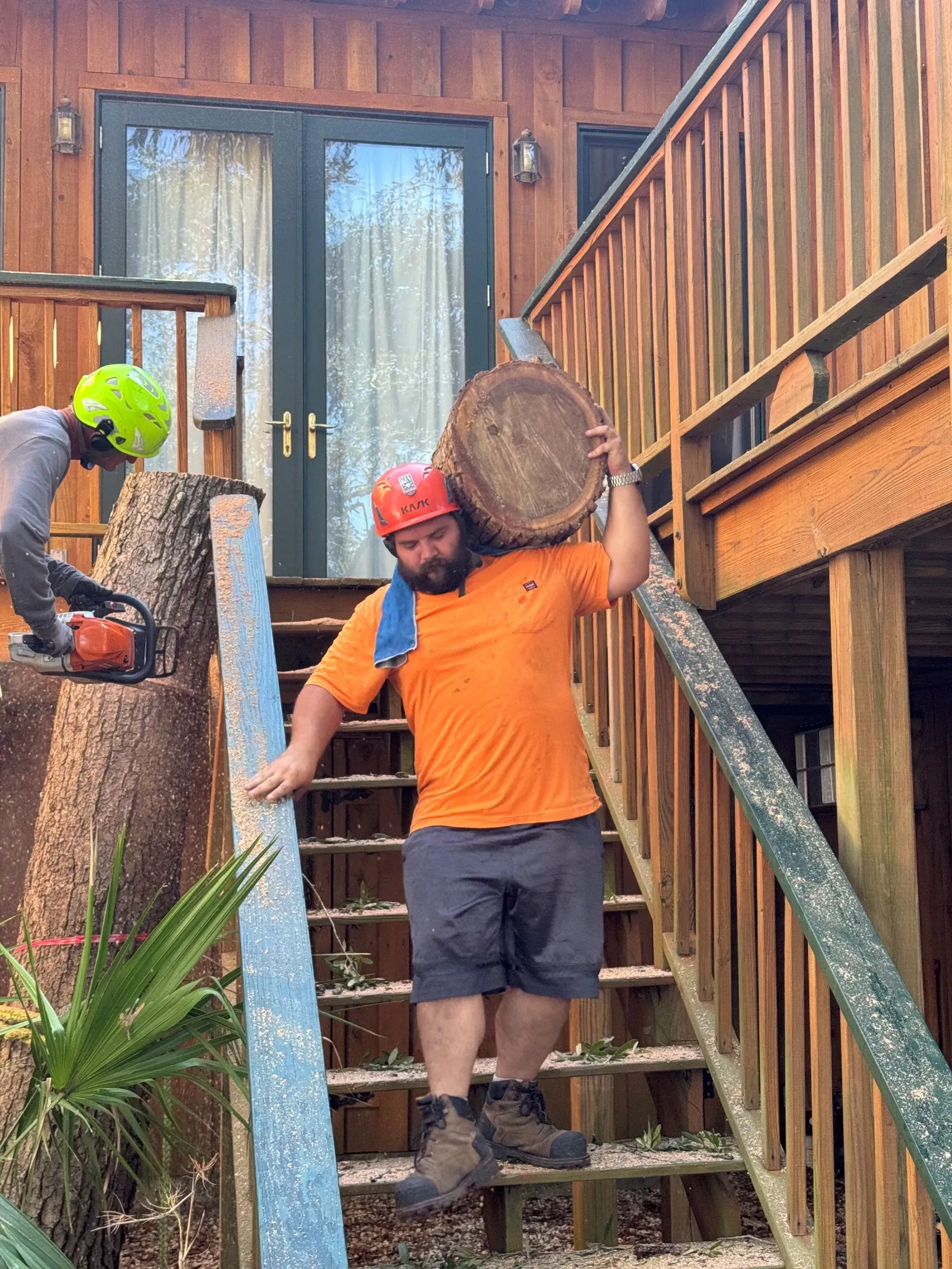 Man carrying a large wooden disc on his shoulder, walking up outdoor stairs. Another person uses a chainsaw.