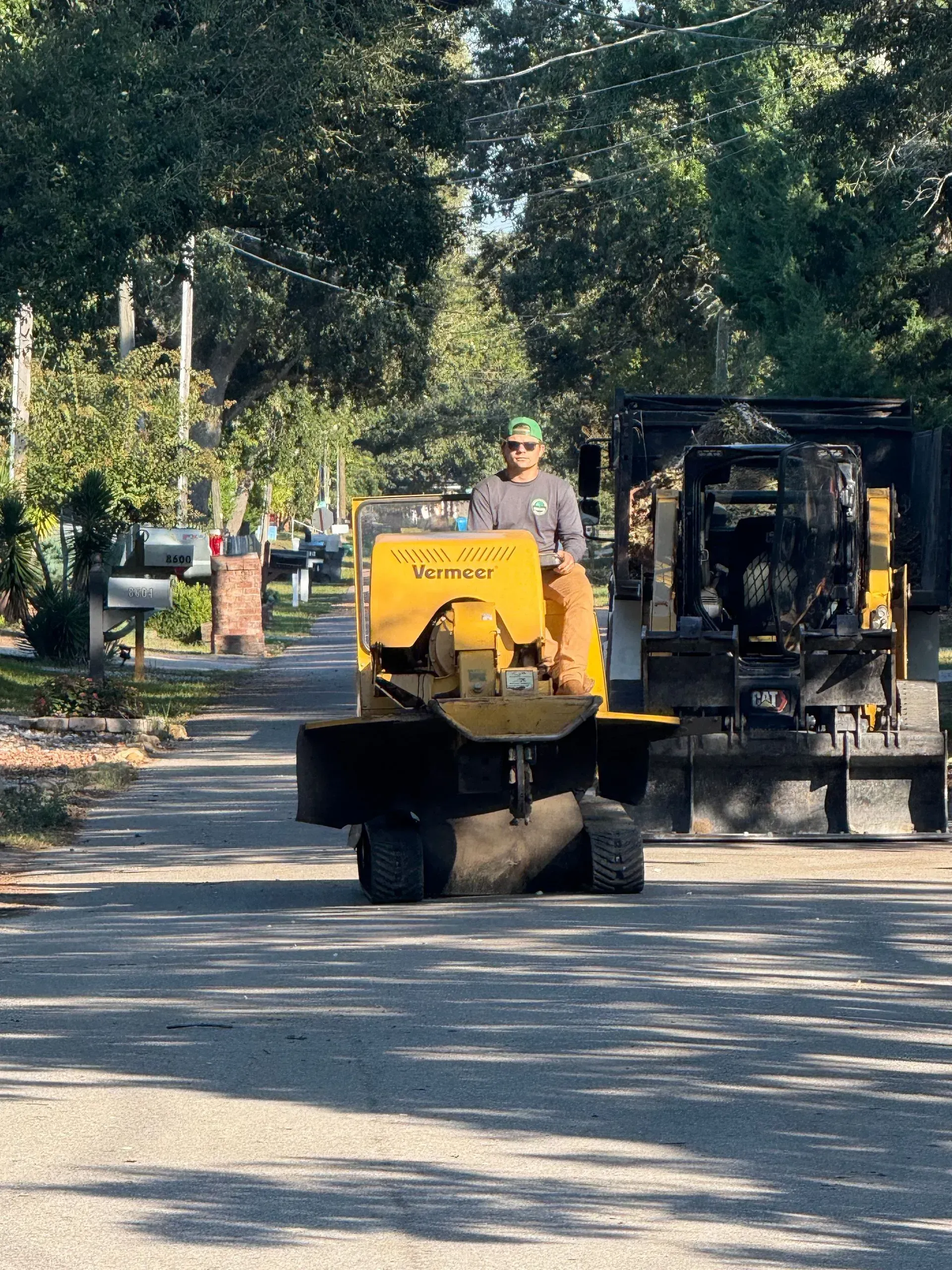 Man operating a yellow stump grinder on a residential street. A trailer is attached to the machine.