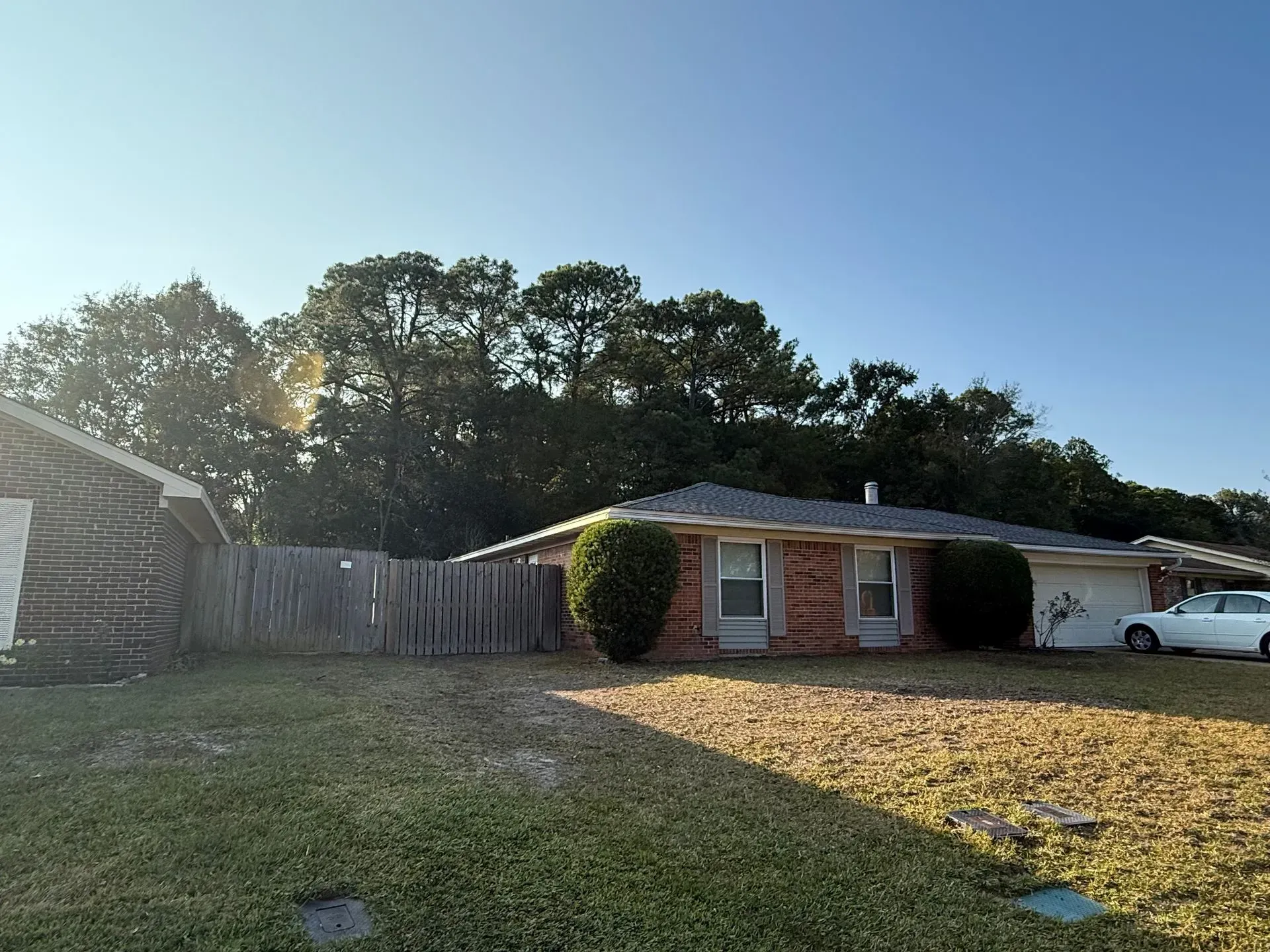 Brick house with brown roof, front yard with grass, wooden fence, trees in background, and a blue sky.