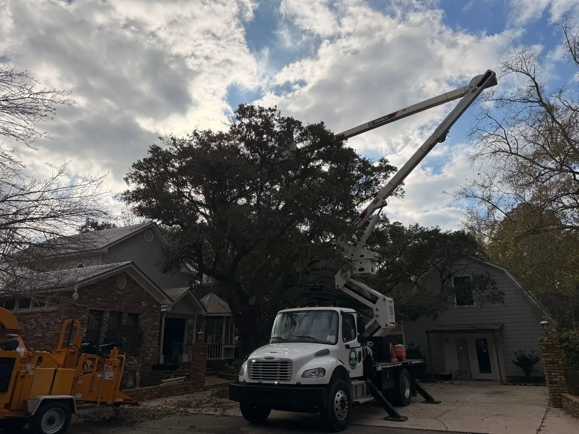 Tree trimming service in progress, truck with extended lift near a house, cloudy sky.