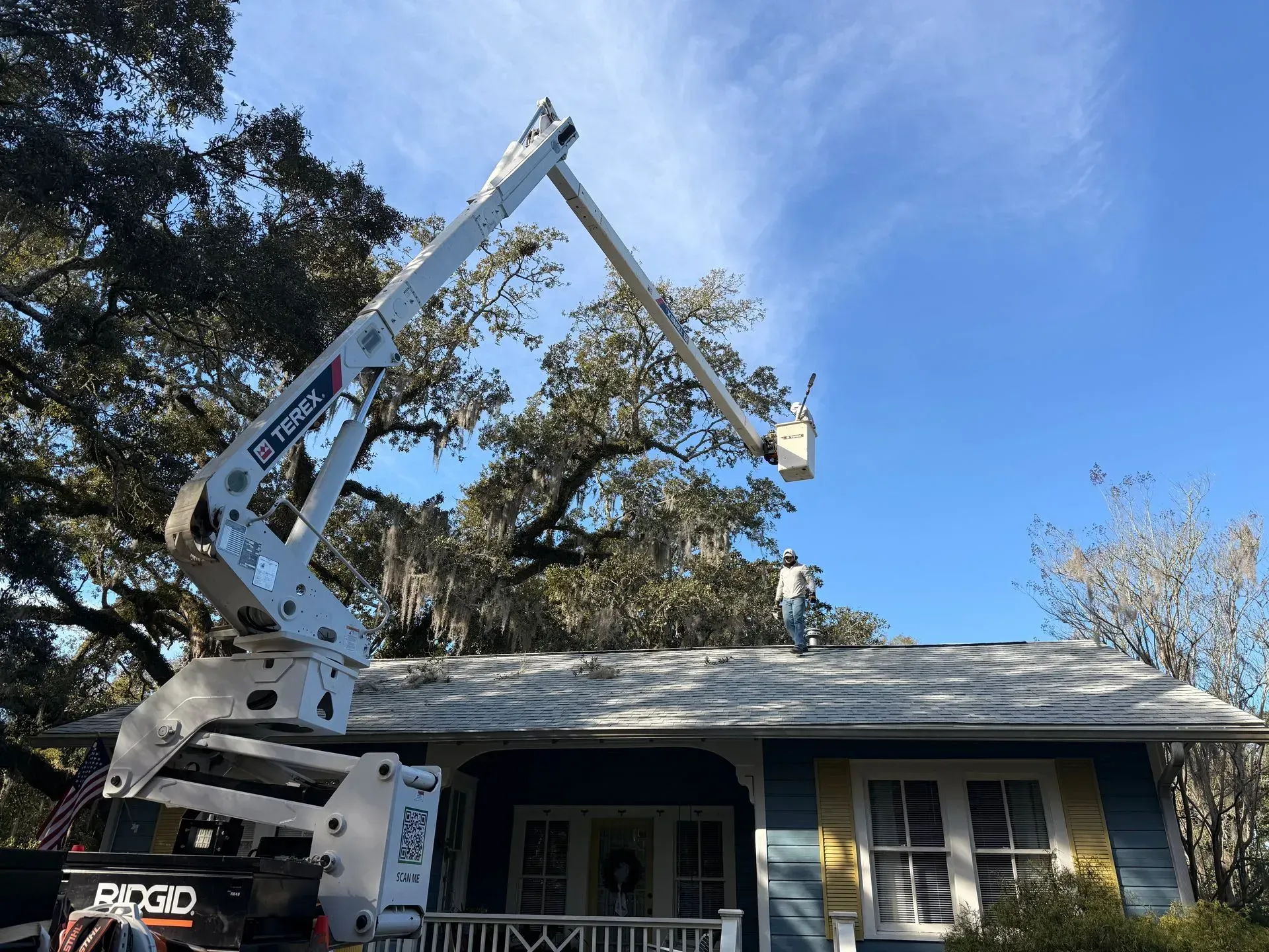 Bucket truck reaching roof of a blue house, worker trimming tree branches on a sunny day.