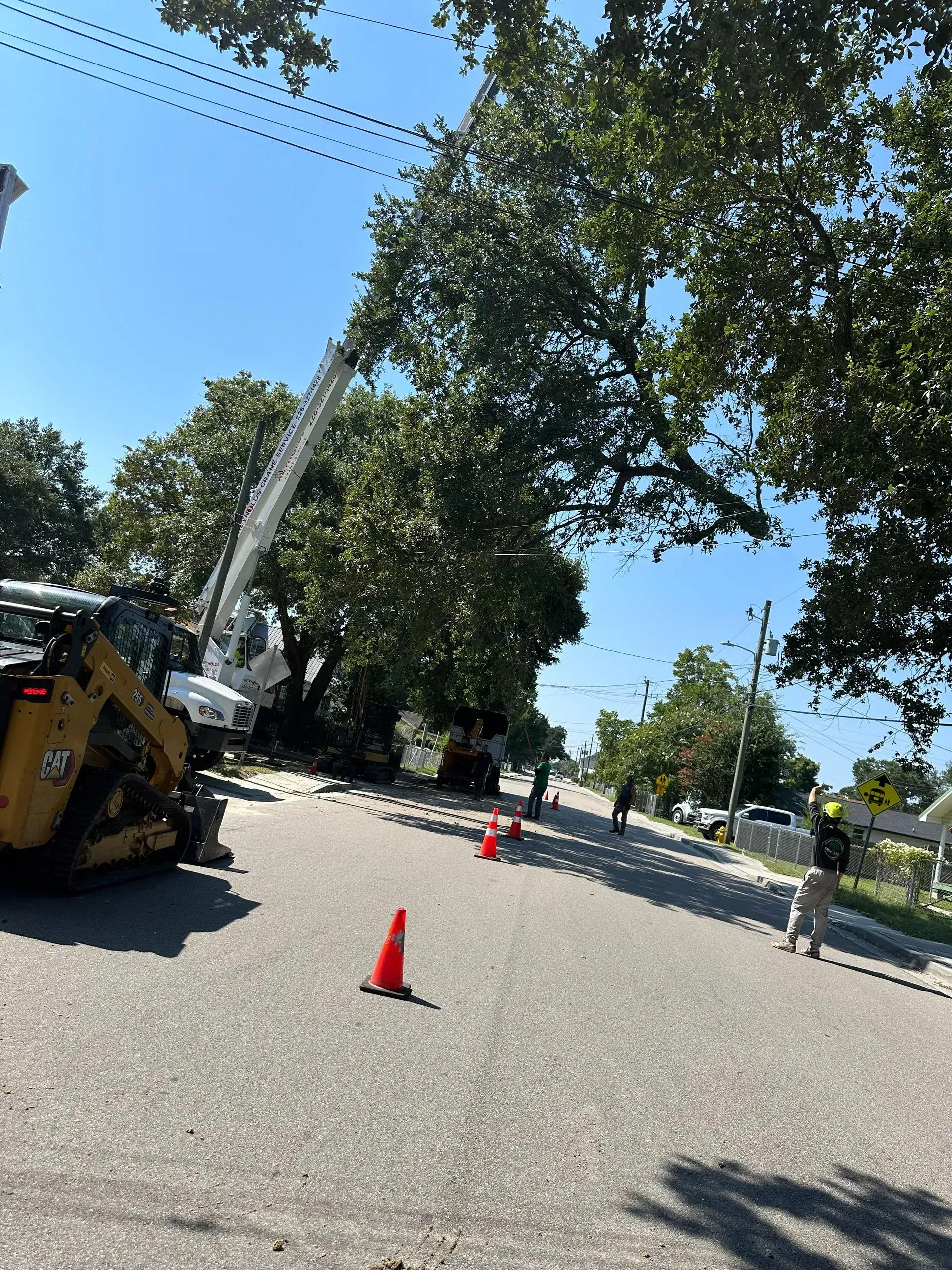 Street scene: Tree trimming by a truck. Workers and orange cones in road. Blue sky.